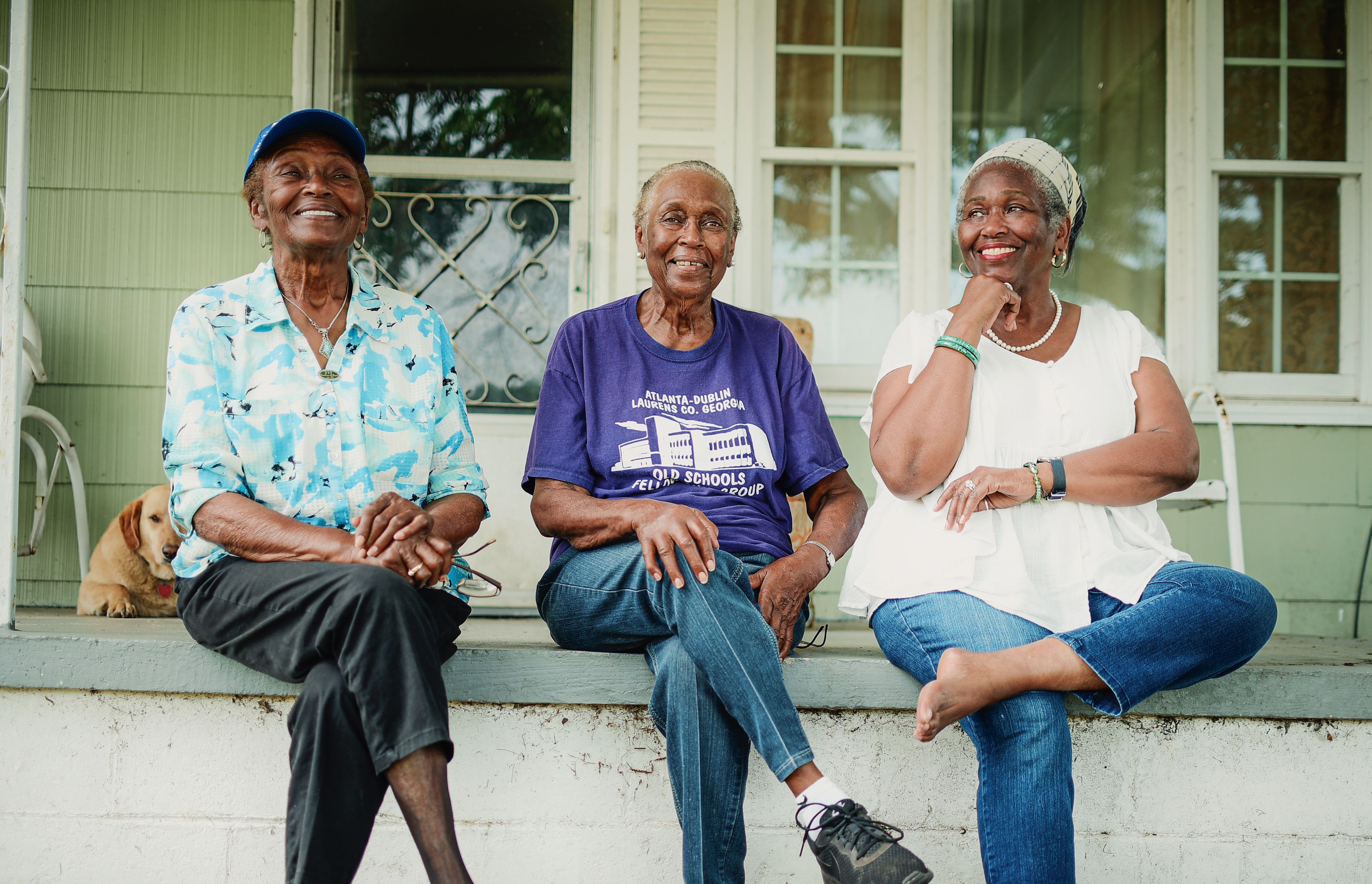 Three sisters sit together, smiling on the edge of their family home's front porch
