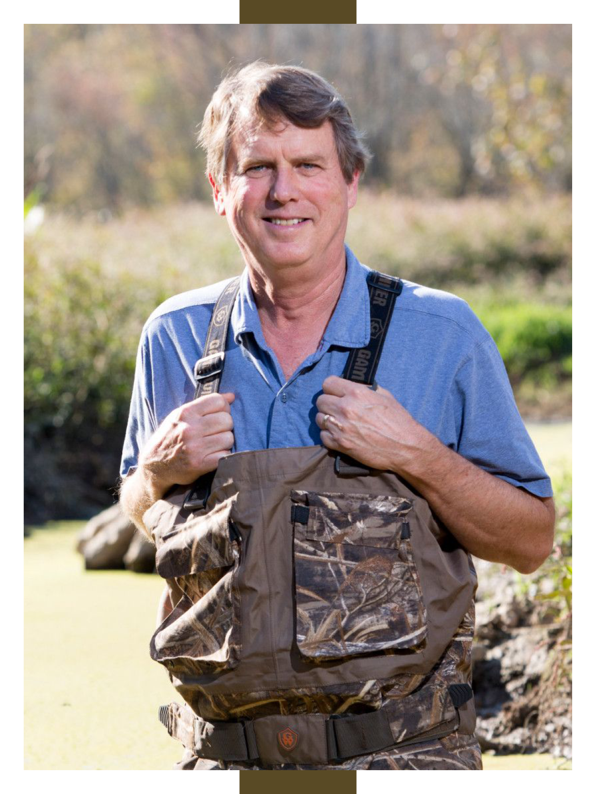 Wearing waders and a blue collared shirt, Darold Batzer stands in a wetland with trees and marsh in the background.