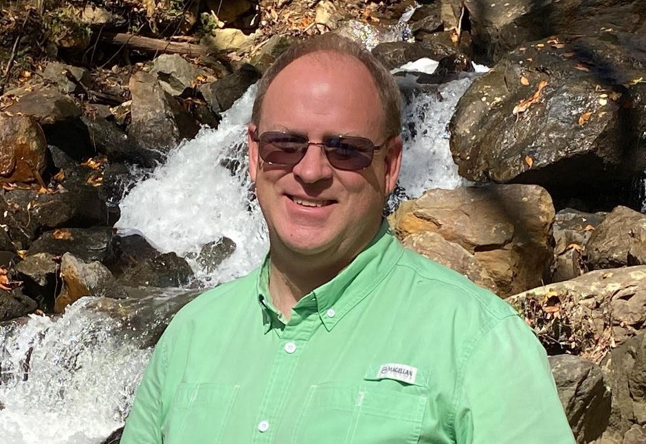 Hawkins smiles while wearing sunglasses and a green shirt while standing in front of a waterfall