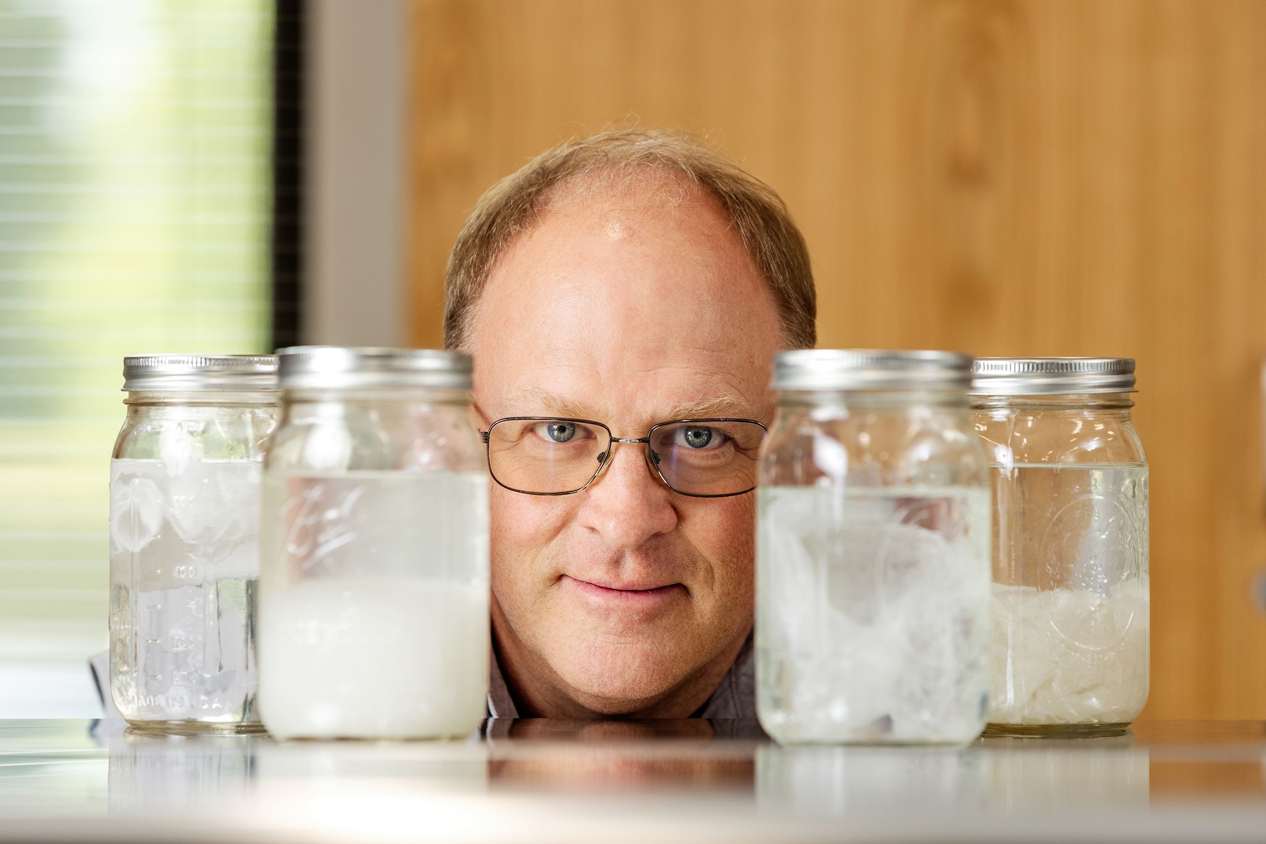 Gary Hawkins poses for a photo with jars containing toilet paper, "flushable" wipes, a dryer sheet, and a paper towel submerged in water. 