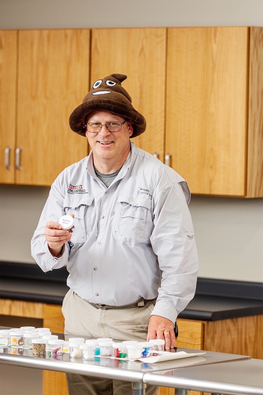 A man in a brown cartoon poop hat stands behind a stainless table bearing small containers