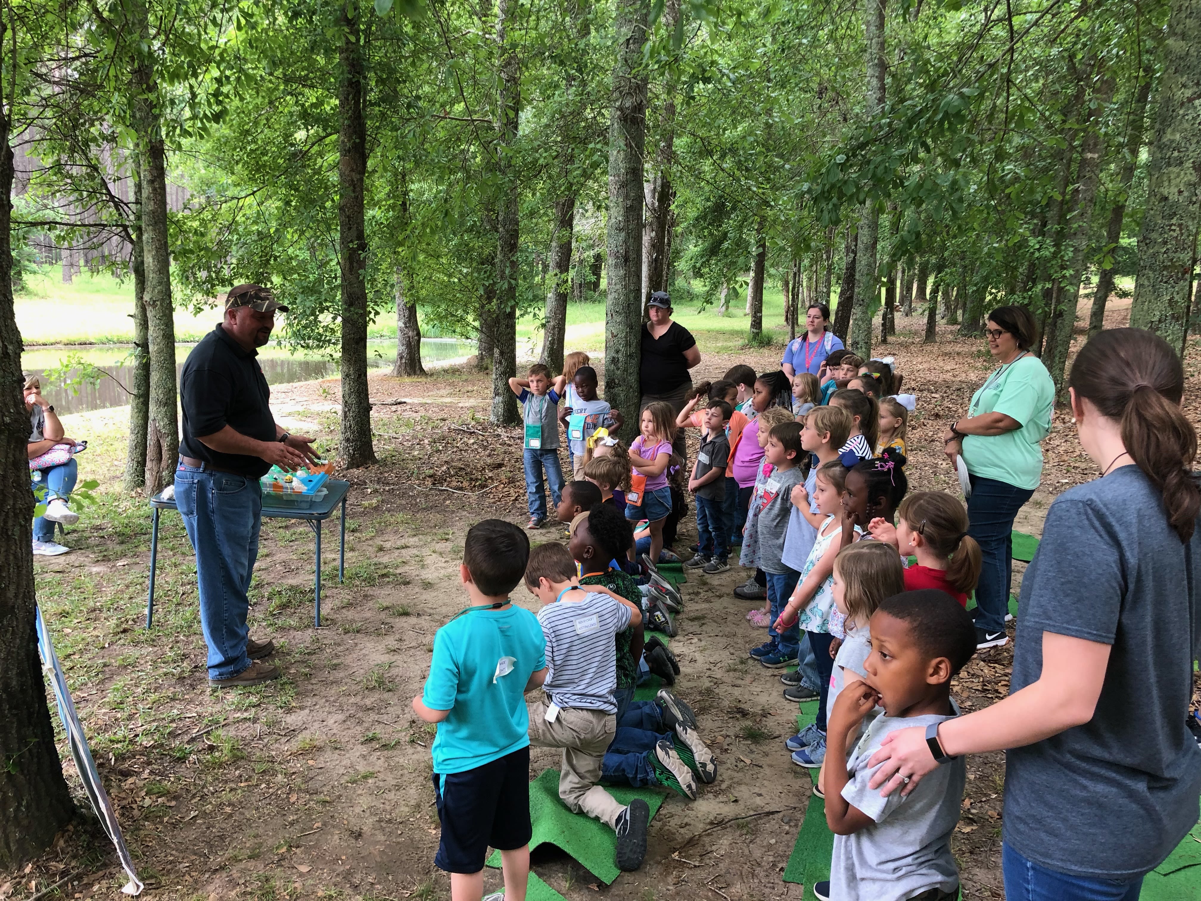 Hall stands in front of a large group of children and their chaperones in a wooded area, where he uses the EnviroScape model on a folding table to demonstrate responsible water use.