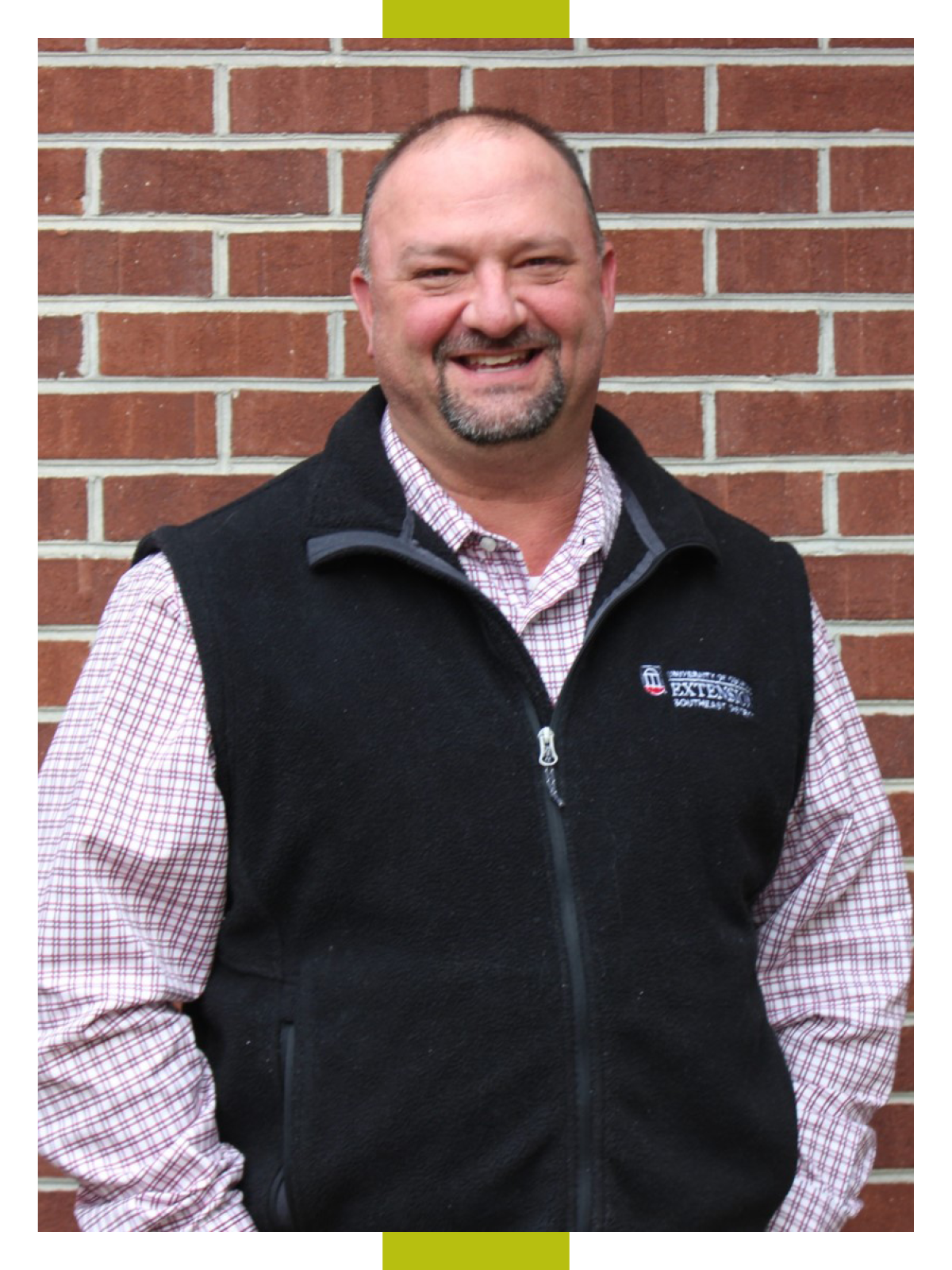 David Hall, smiling, stands in front of a brick wall wearing a plaid short and black UGA Extension branded fleece vest
