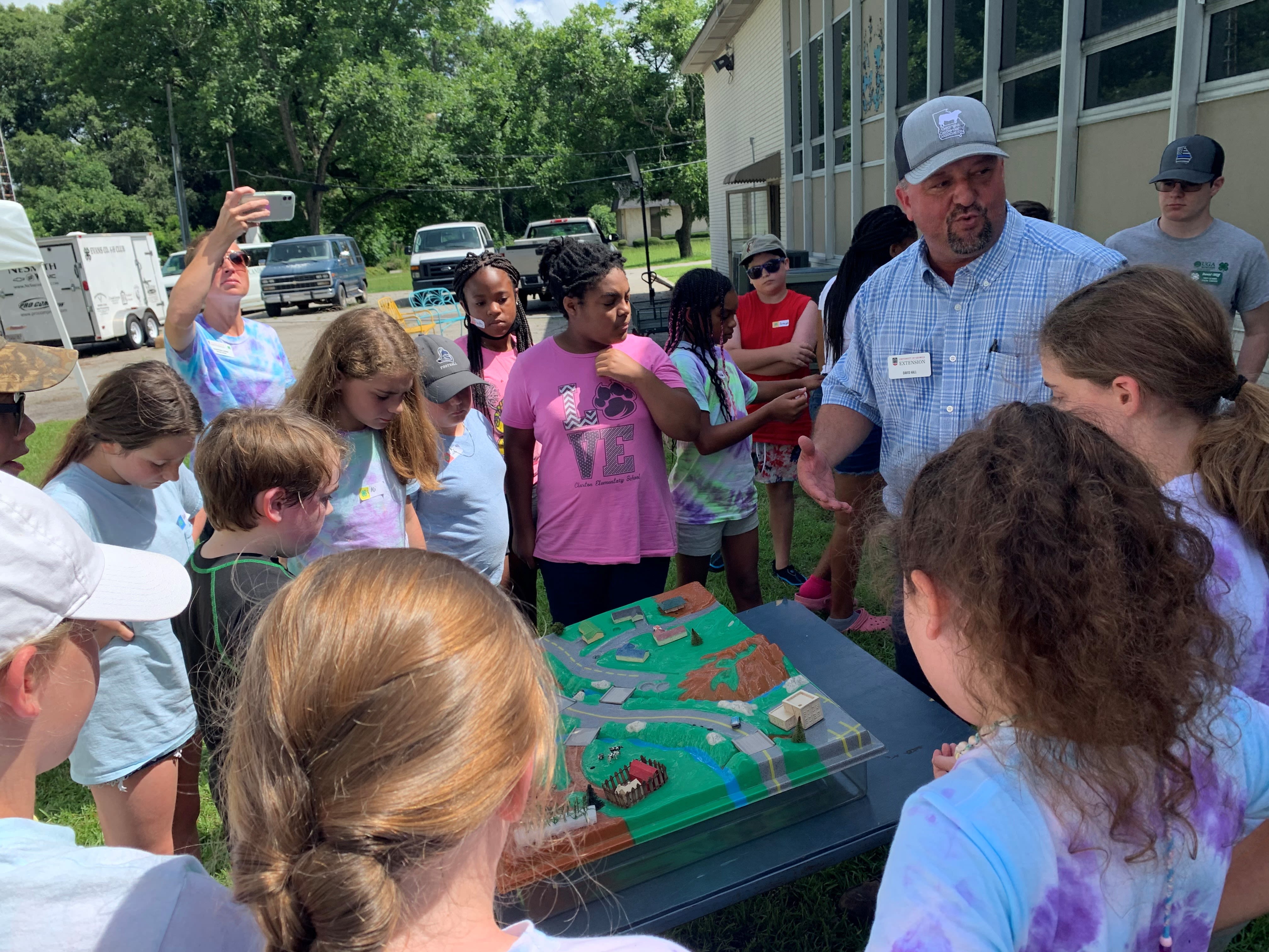 A group of yound students gathers around Hall and the EnviroScape model in a parking lot where they are learning about water conservation.