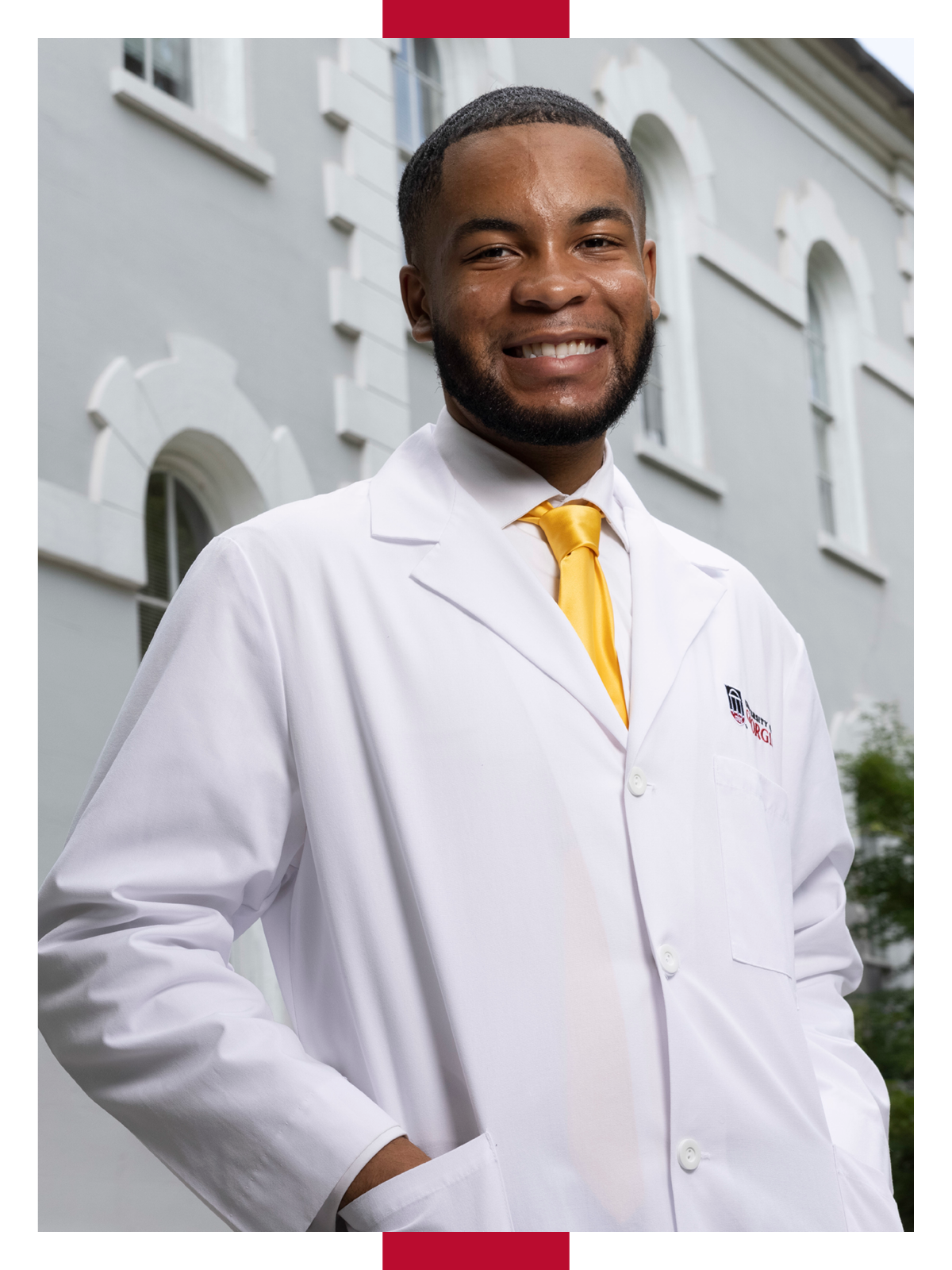 Eric Okanume wears a white lab coat with a UGA logo and a yellow tie and smiles while standing on UGA's North Campus