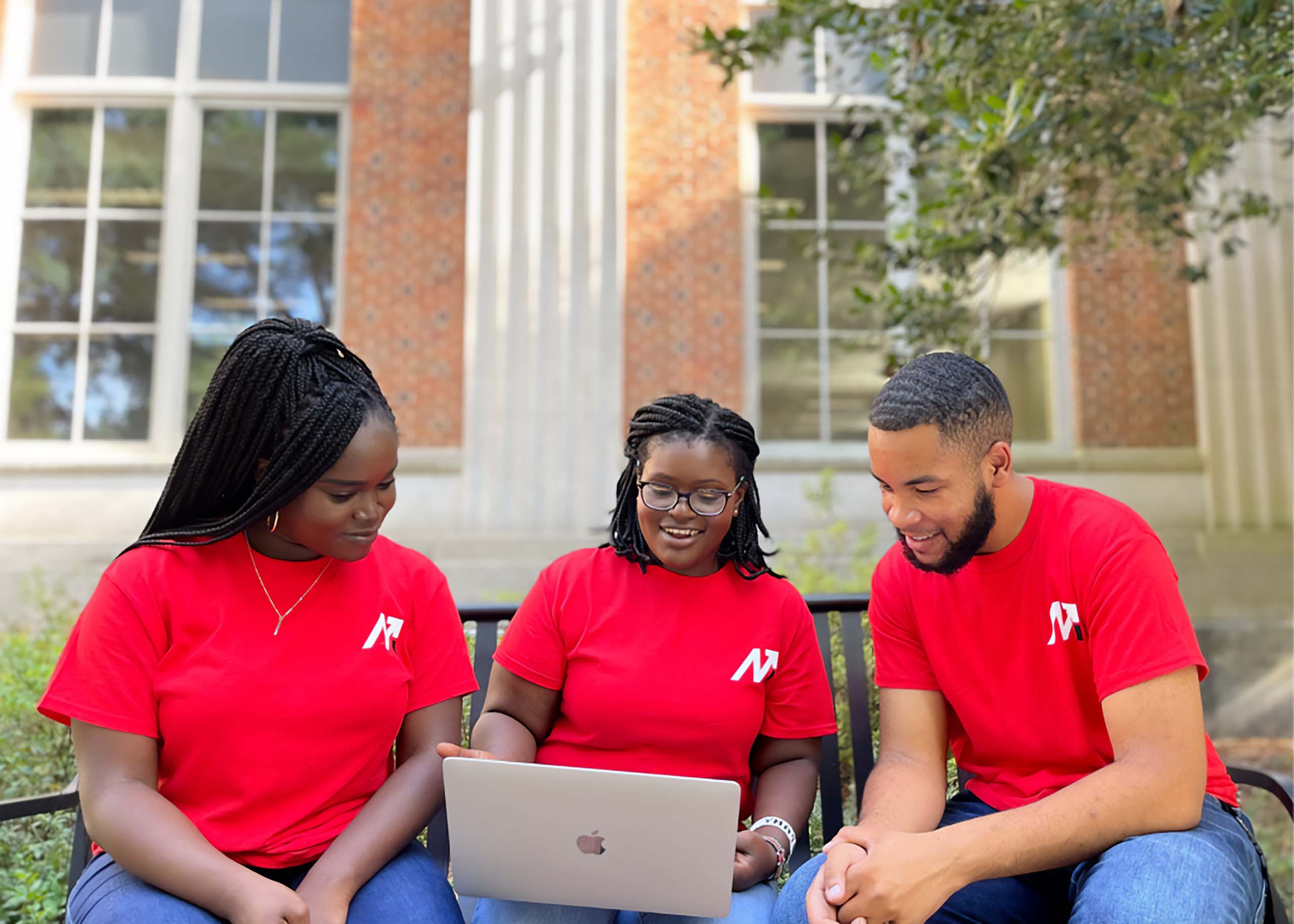 Three students in red shirts gaze at a laptop while sitting outside on campus
