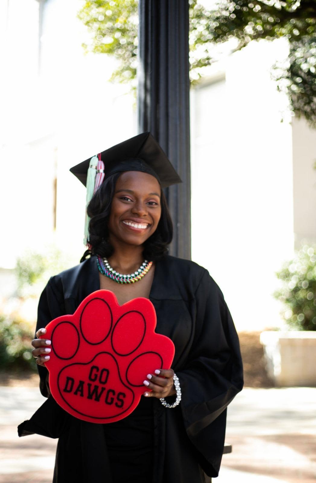 Phyllicia Thomas in a black cap and gown holds a paw print shaped foam sign proclaiming Go Dawgs at her UGA graduation in 2019. 