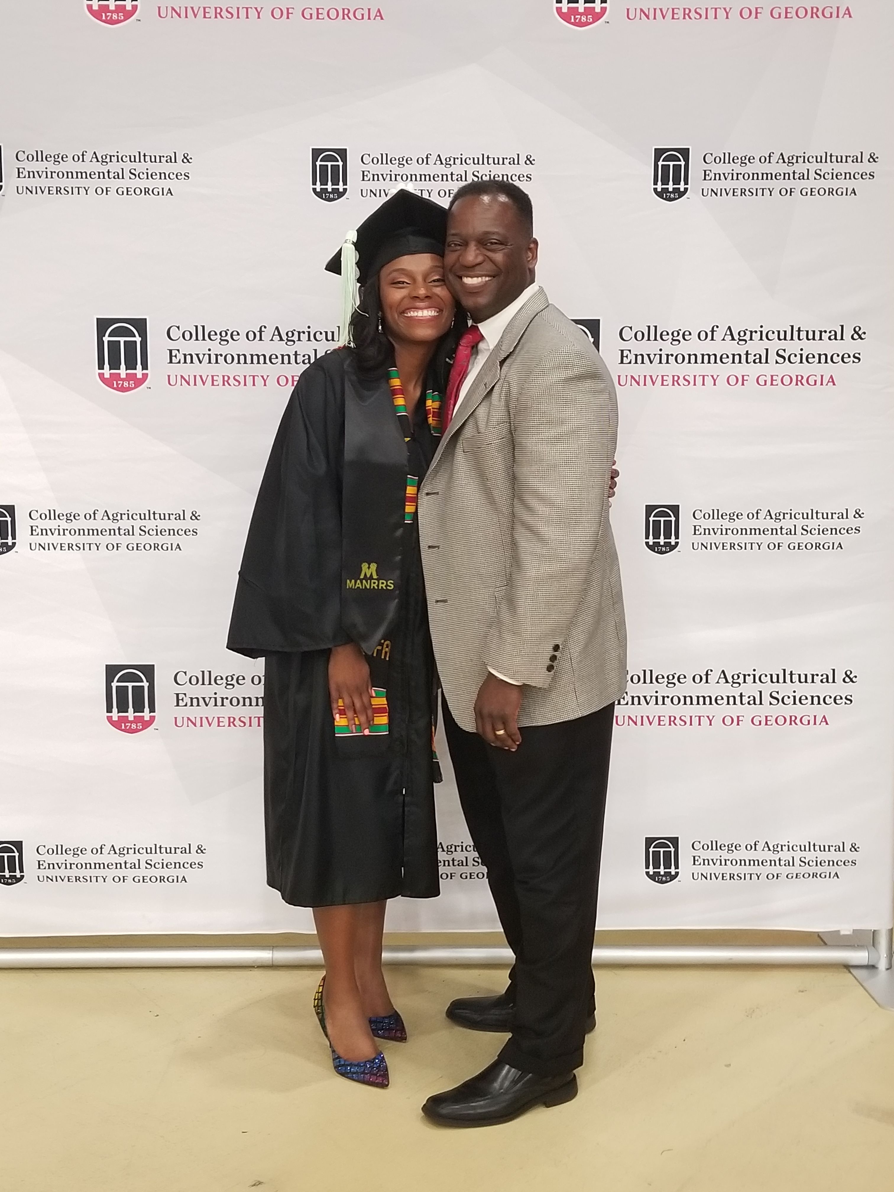 Phyllicia Thomas, in a black cap and gown, and her father, Chris Thomas, in a suit stand in front of a CAES logo backdrop