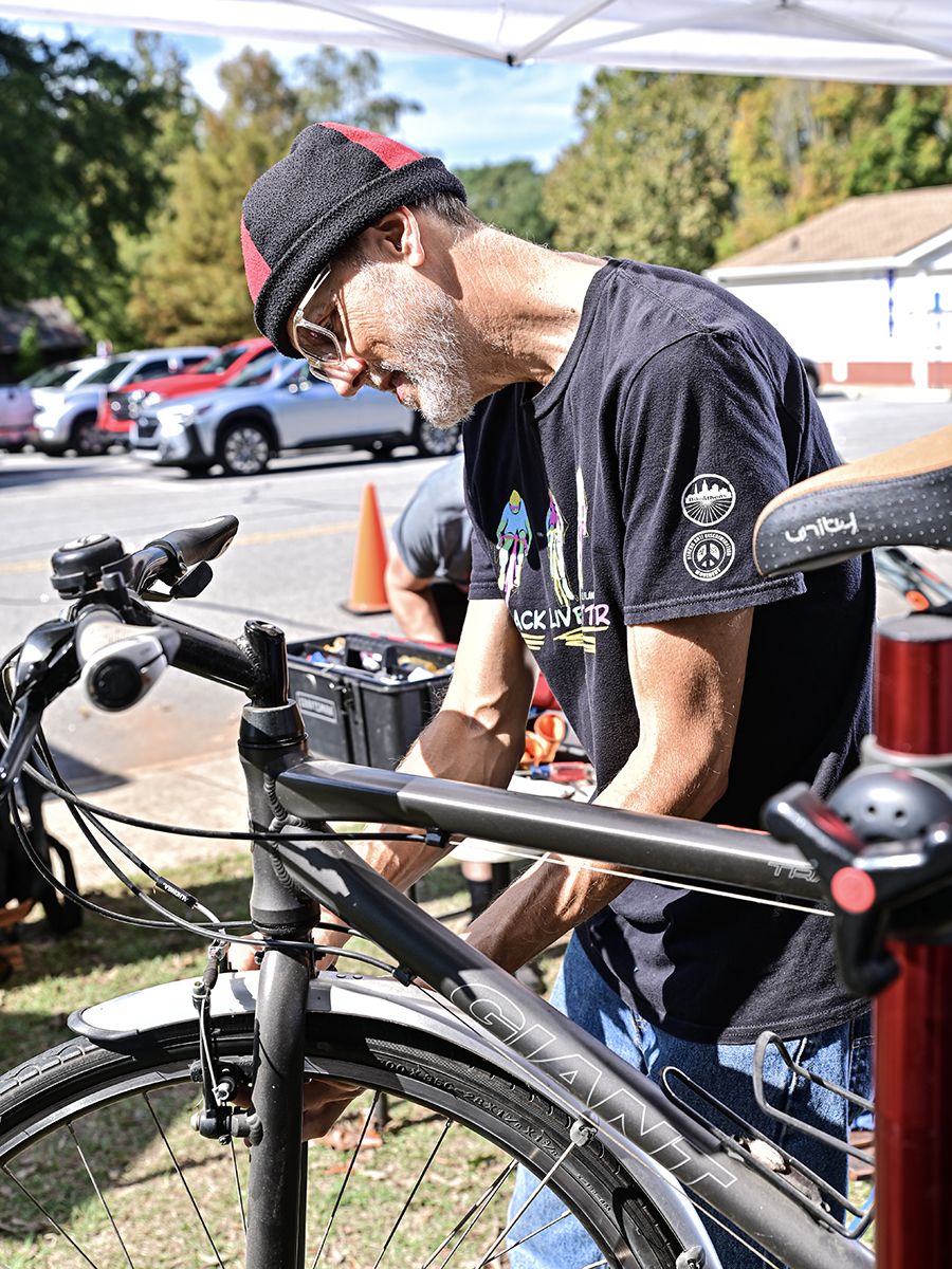 A man wearing a beanie and glasses repairs the front wheel of a black bicycle at an outdoor event, with tools and cars visible in the background.