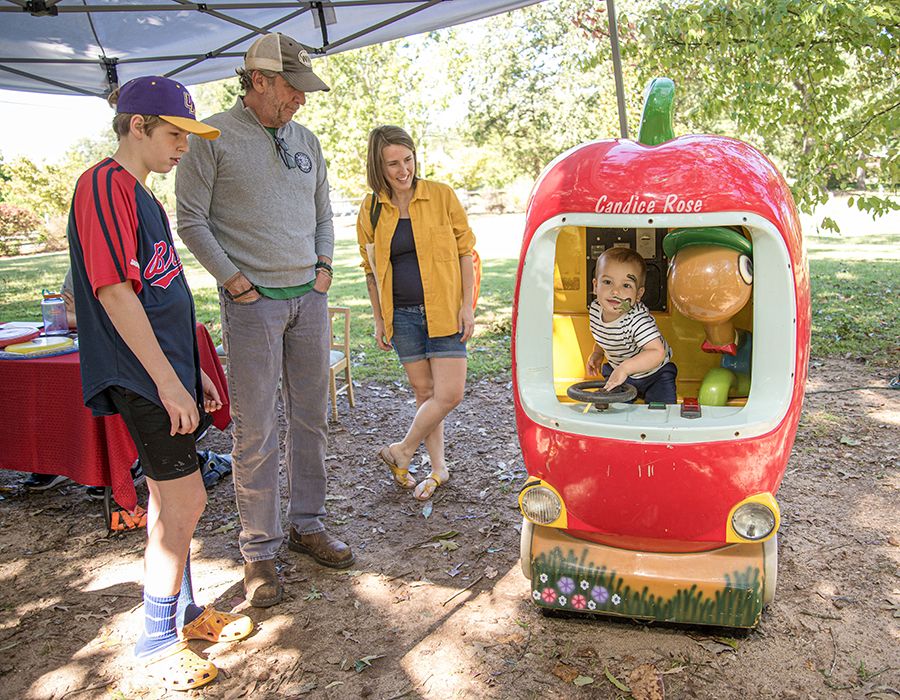 A smiling toddler sits inside an apple-shaped ride-on vehicle while a man, woman and teenager watch nearby under a canopy in a park setting.     