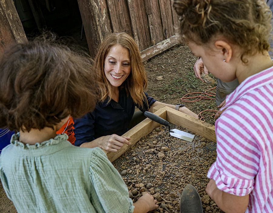 A woman smiles as she helps children sift through dirt and rocks using a wooden screen during an outdoor educational activity near a rustic wooden building.