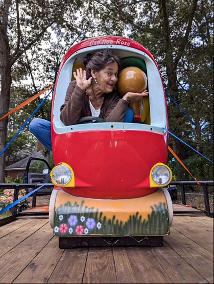 A woman playfully poses and waves from inside a colorful, apple-shaped ride-on vehicle set on a wooden platform outdoors.
