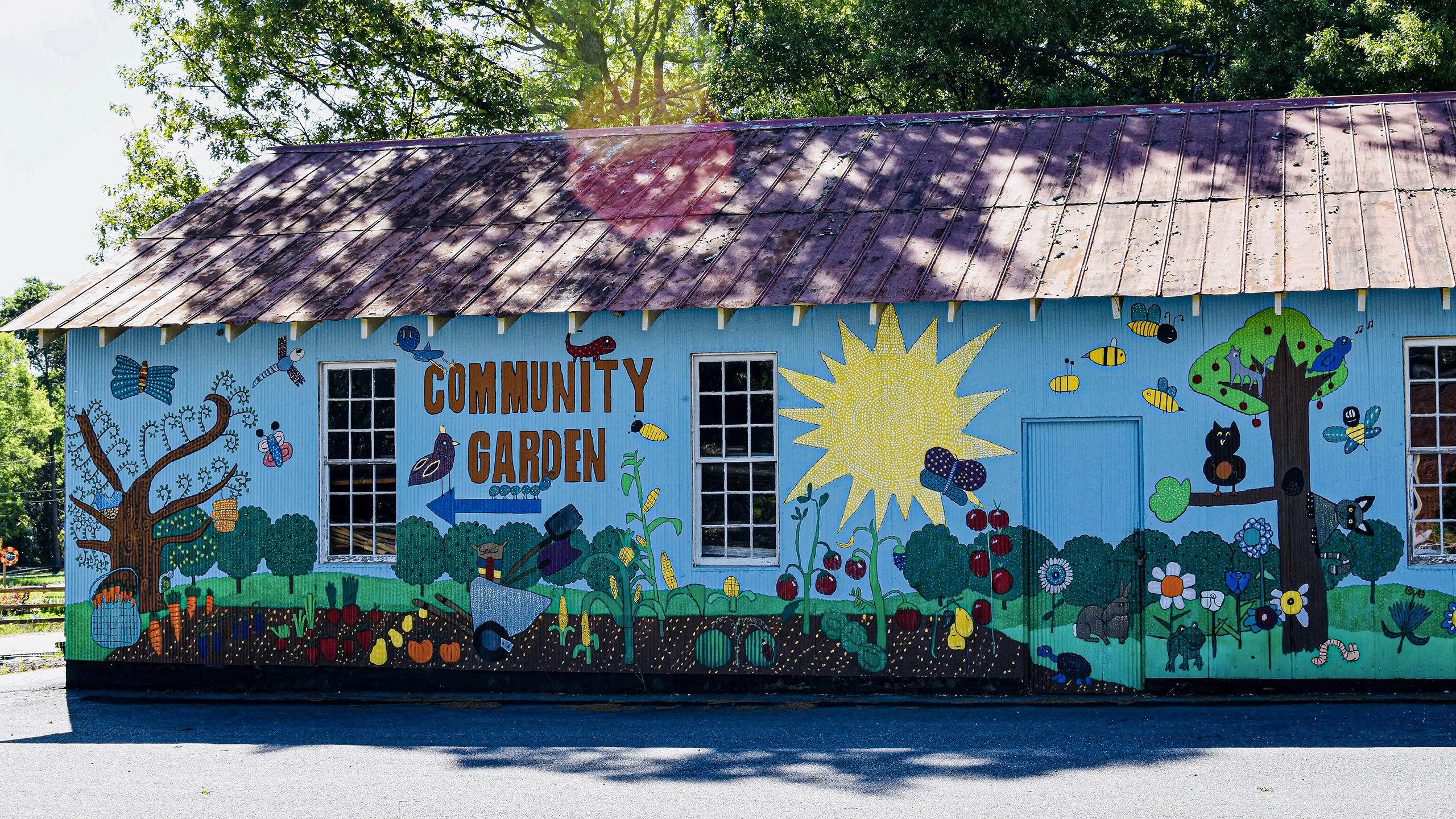 A mural on the side of a one story building features a sky-blue background and large colorful illustrations of a vegetable garden, whimsical trees and wildlife.