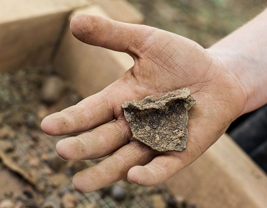 A dirt-covered hand holds a rough, triangular piece of pottery or stone, with a wooden sifting screen and scattered debris in the background.