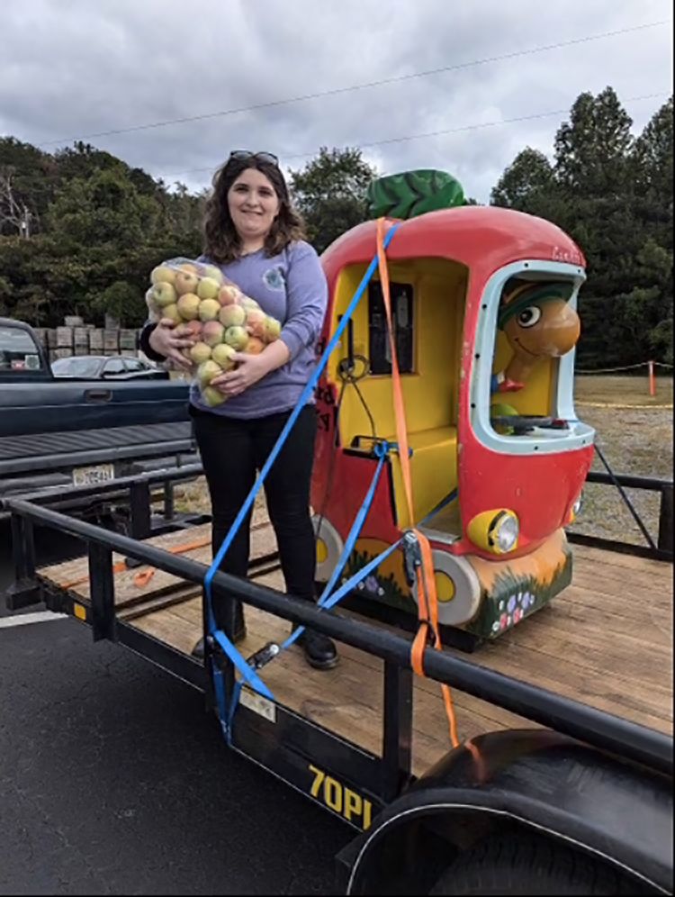 A woman stands on a trailer holding a bundle of apples beside a colorful, apple-shaped ride-on toy car.