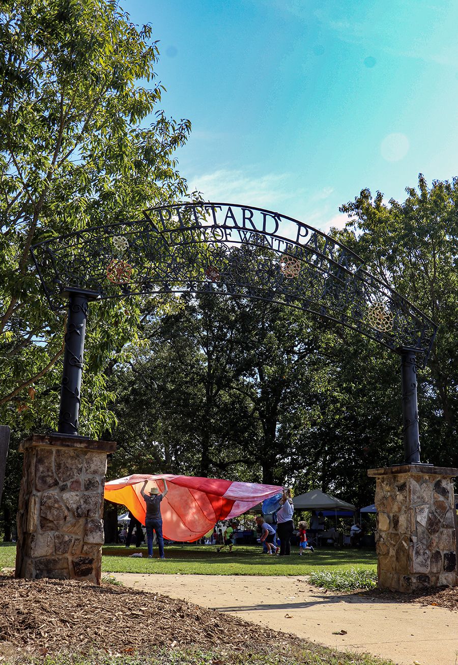 People play with a colorful parachute under the decorative iron entrance arch of Pittard Park on a sunny day, surrounded by trees and vendor tents.