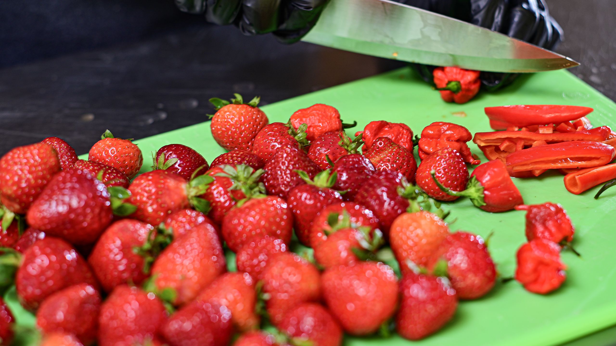 A large pile of bright red strawberries sit on a green cutting board on which a knife is shown slicing small red peppers.