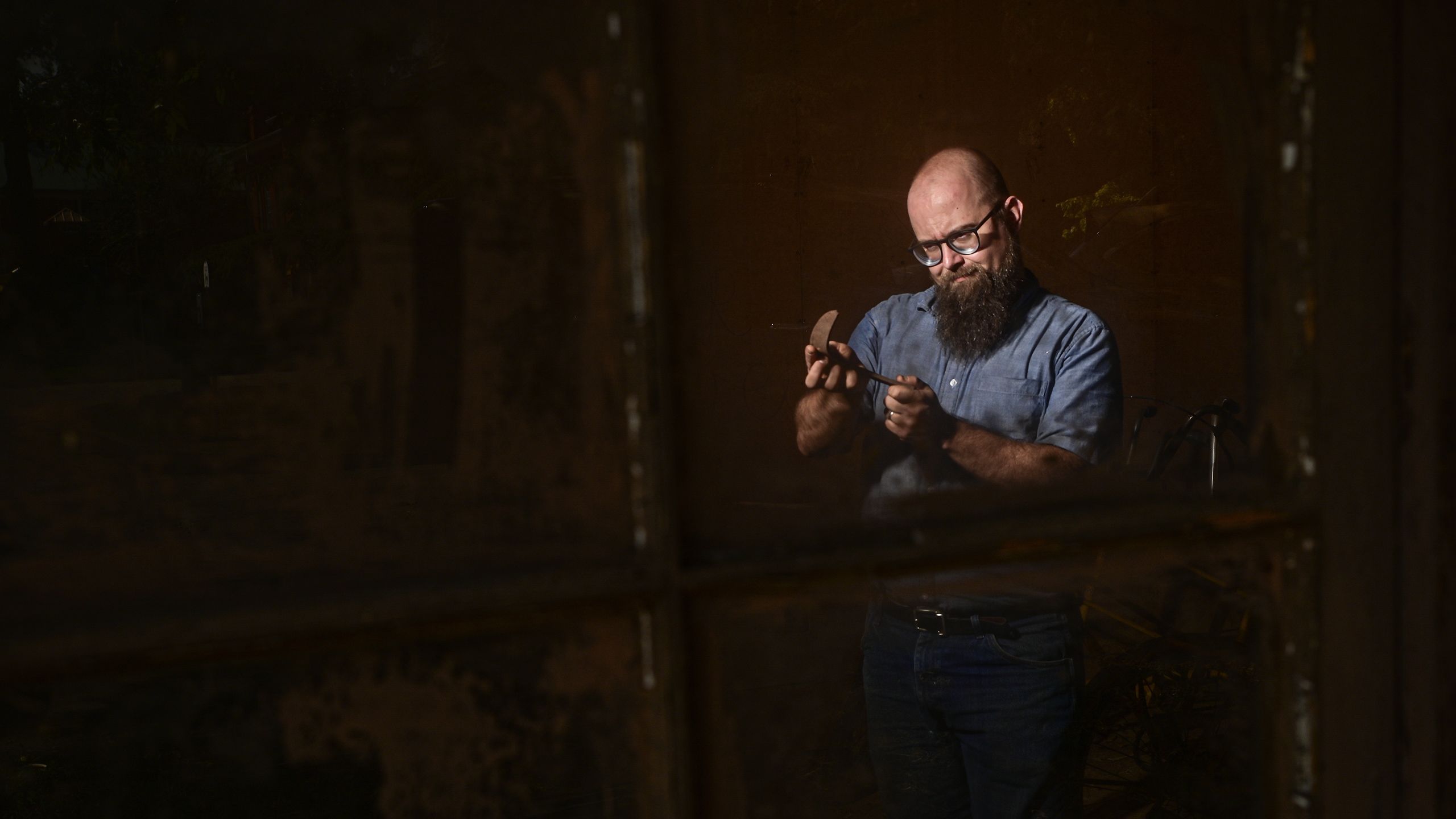 A bearded man wearing glasses and a denim shirt carefully inspects or shapes a curved metal tool under focused lighting.