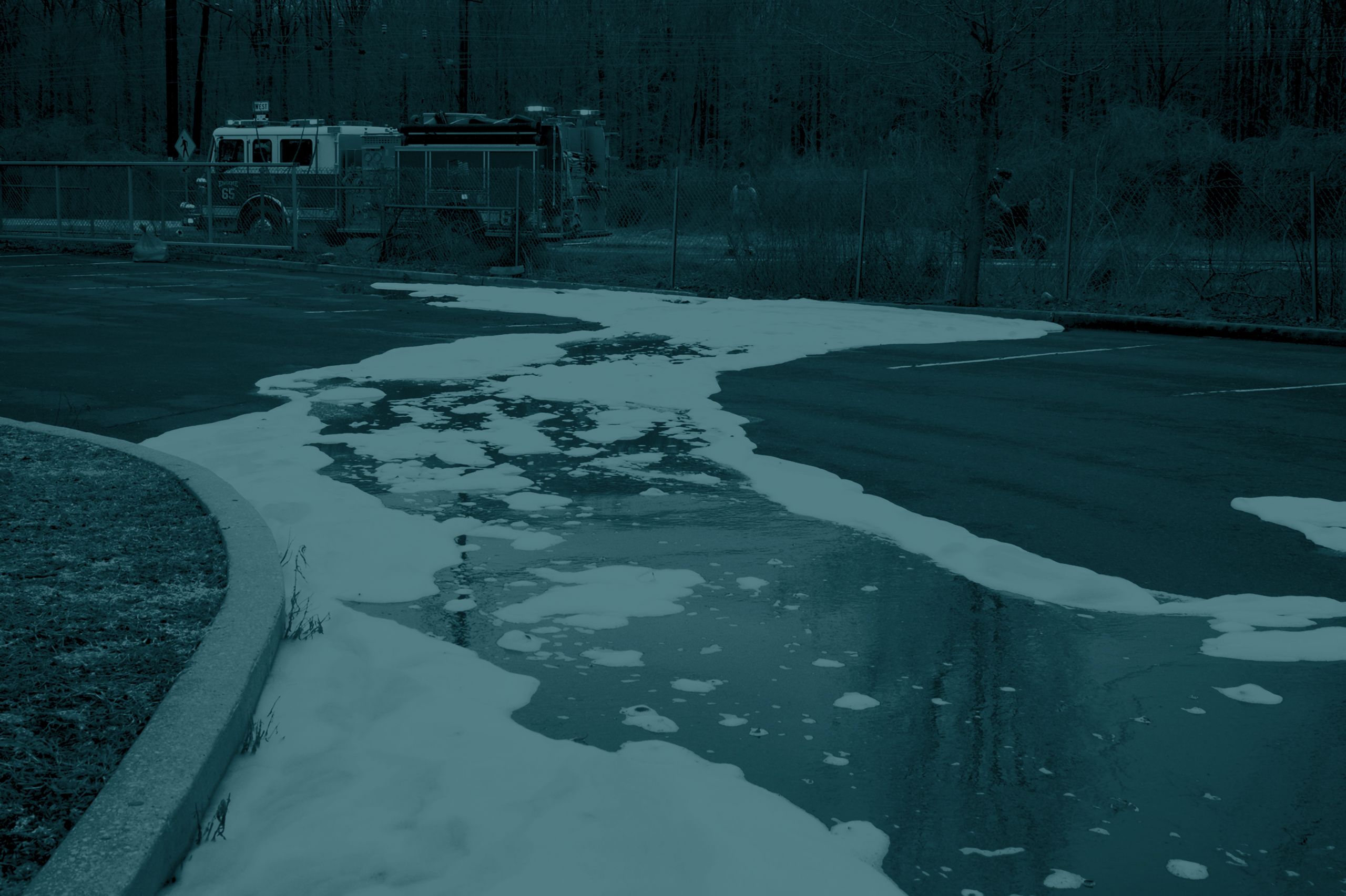 A foamy substance spills onto a paved area, with fire trucks visible in the background.