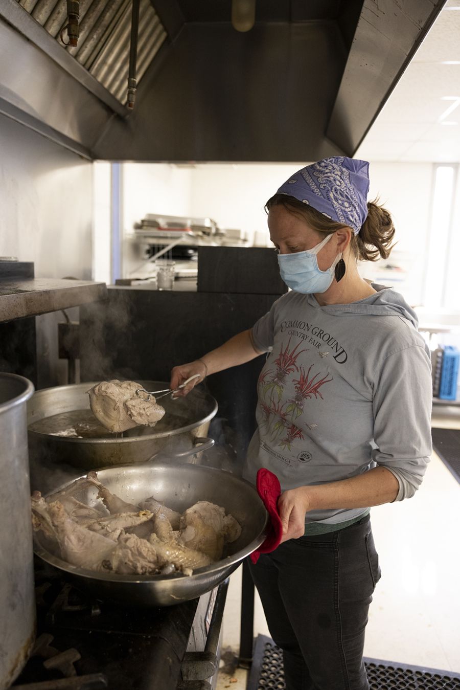 Andie Bisceglia, wearing a grey sweatshirt and a purple bandana on her head, pulls turkey from a pot as she cooks in an industrial kitchen.