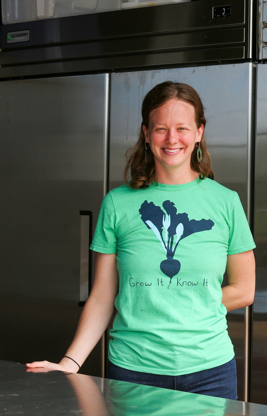 Andie Bisceglia standing in an industrial kitchen. Andie is wearing a green shirt that says "grow it know it" and smiling at the camera.