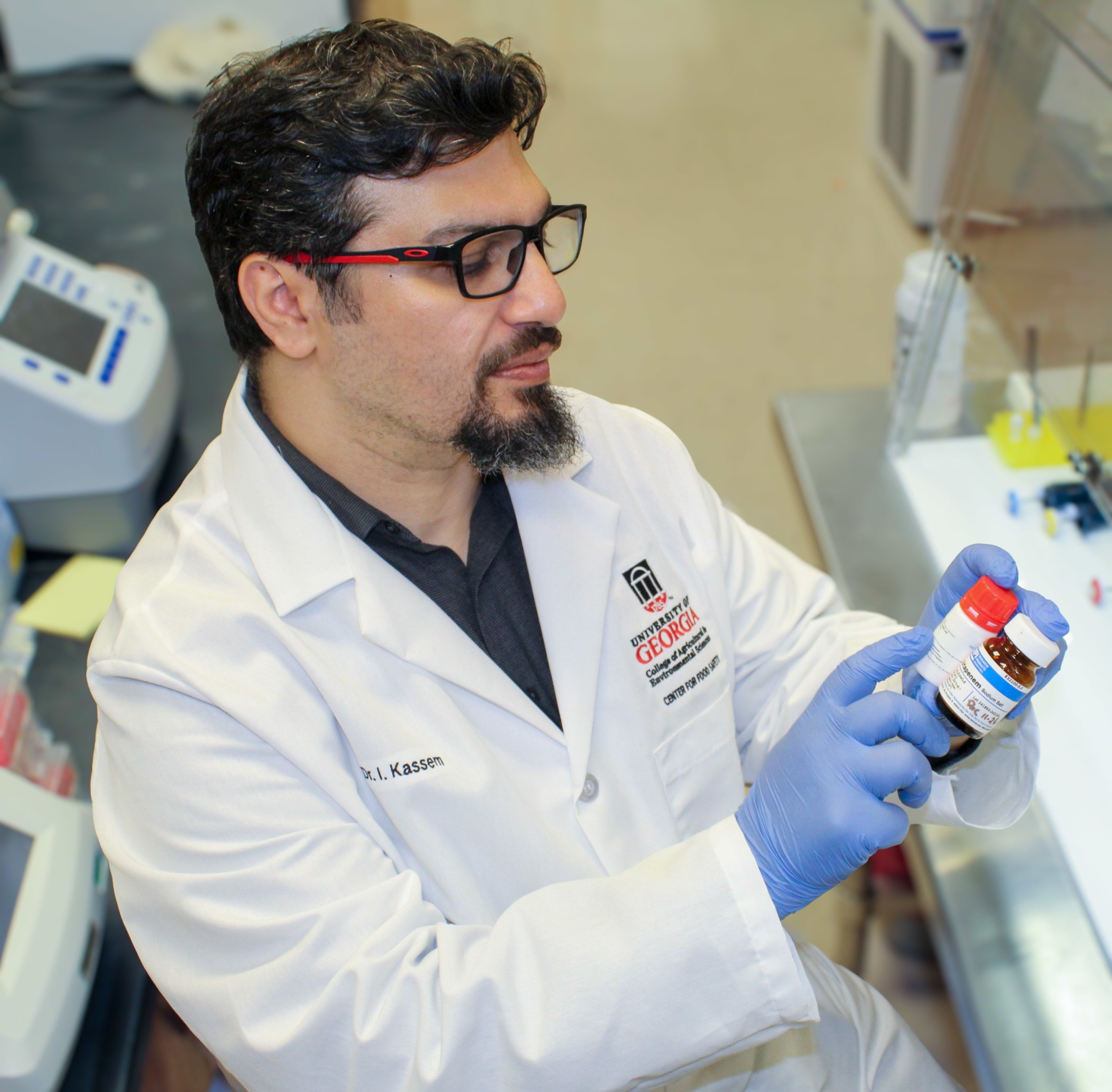 Issmat Kassem looks at two small bottles while wearing a white lab coat and blue gloves in a lab sestting.