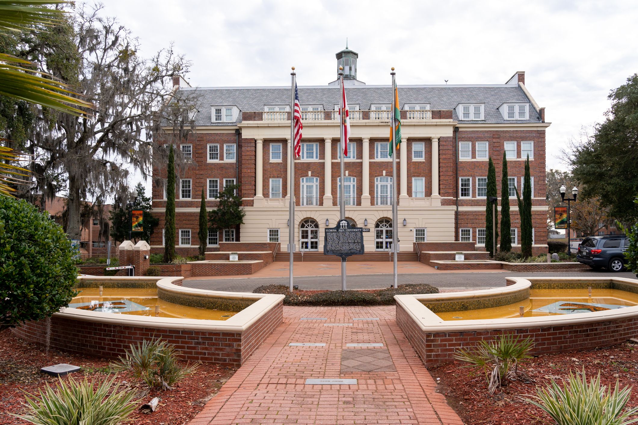 Wide photo of a brick terrace and three flagpoles in front of a brick higher ed building