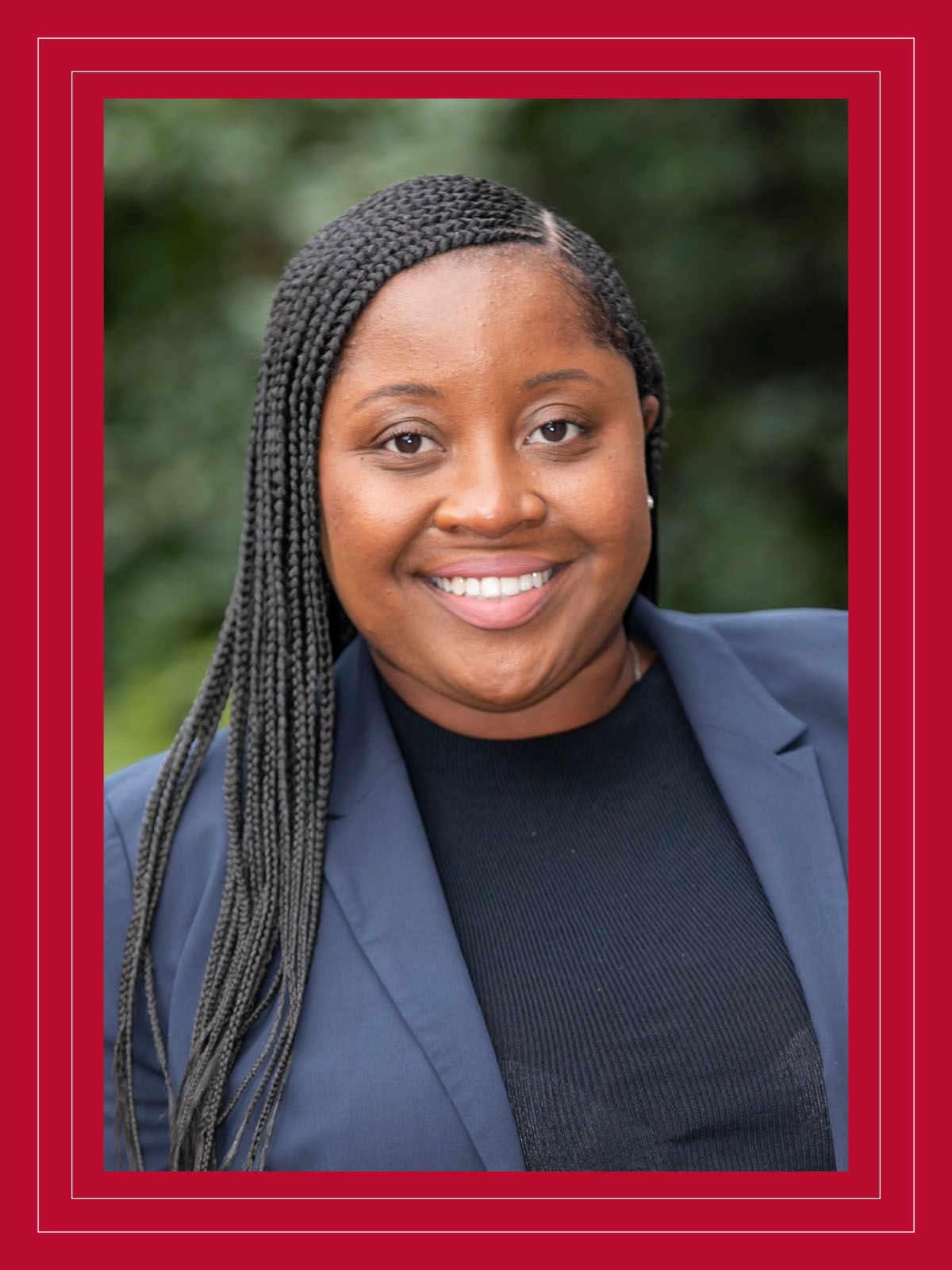 Smiling agent in blue blazer and black shirt standing in front of trees and shrubs.