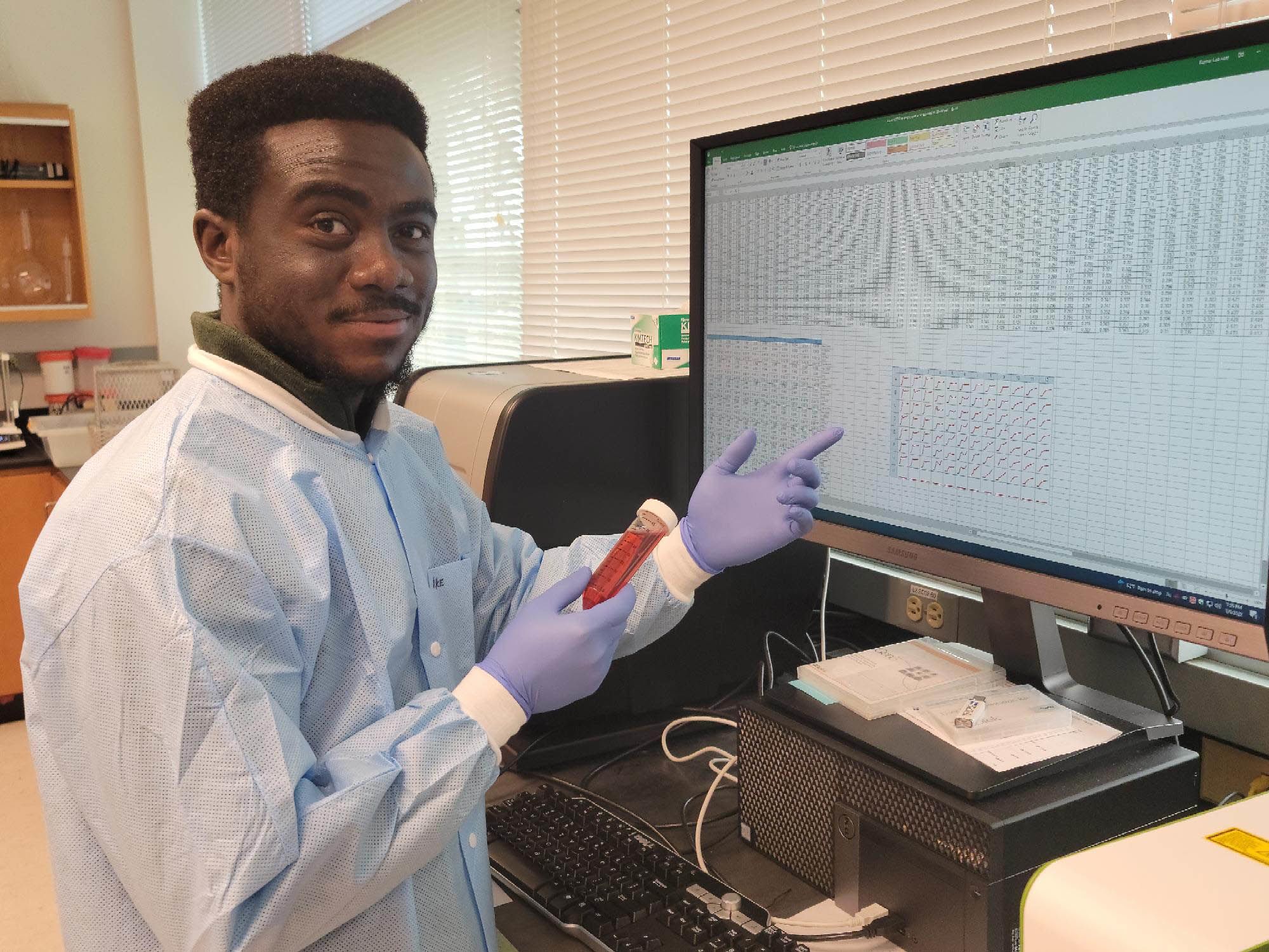Man wearing a lab coat and gloves holds a vial of roselle extract in front of a monitor in a lab