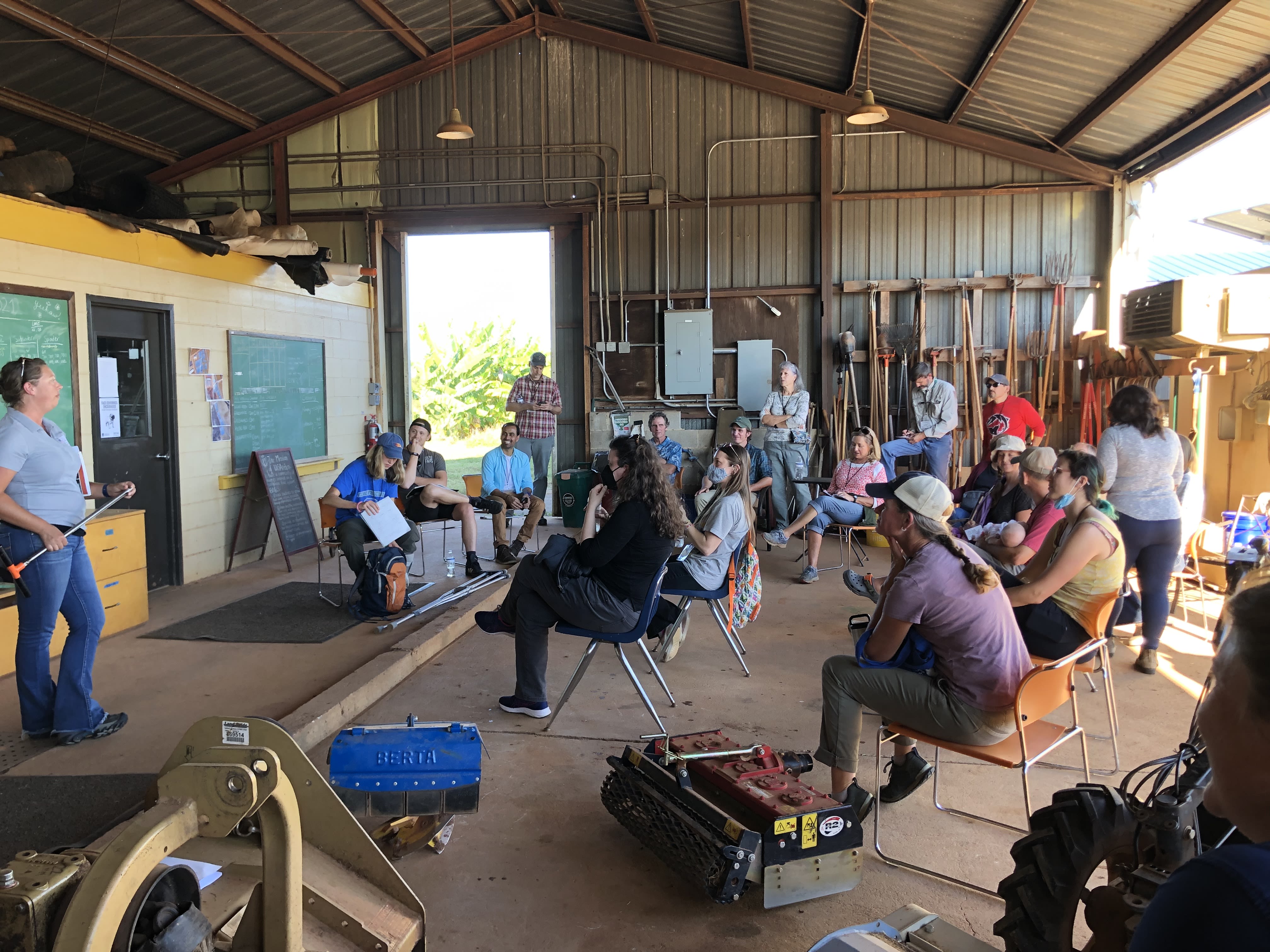 Partipants set in plastic chairs in a large teaching barn equipped with blackboards as well as farming tools