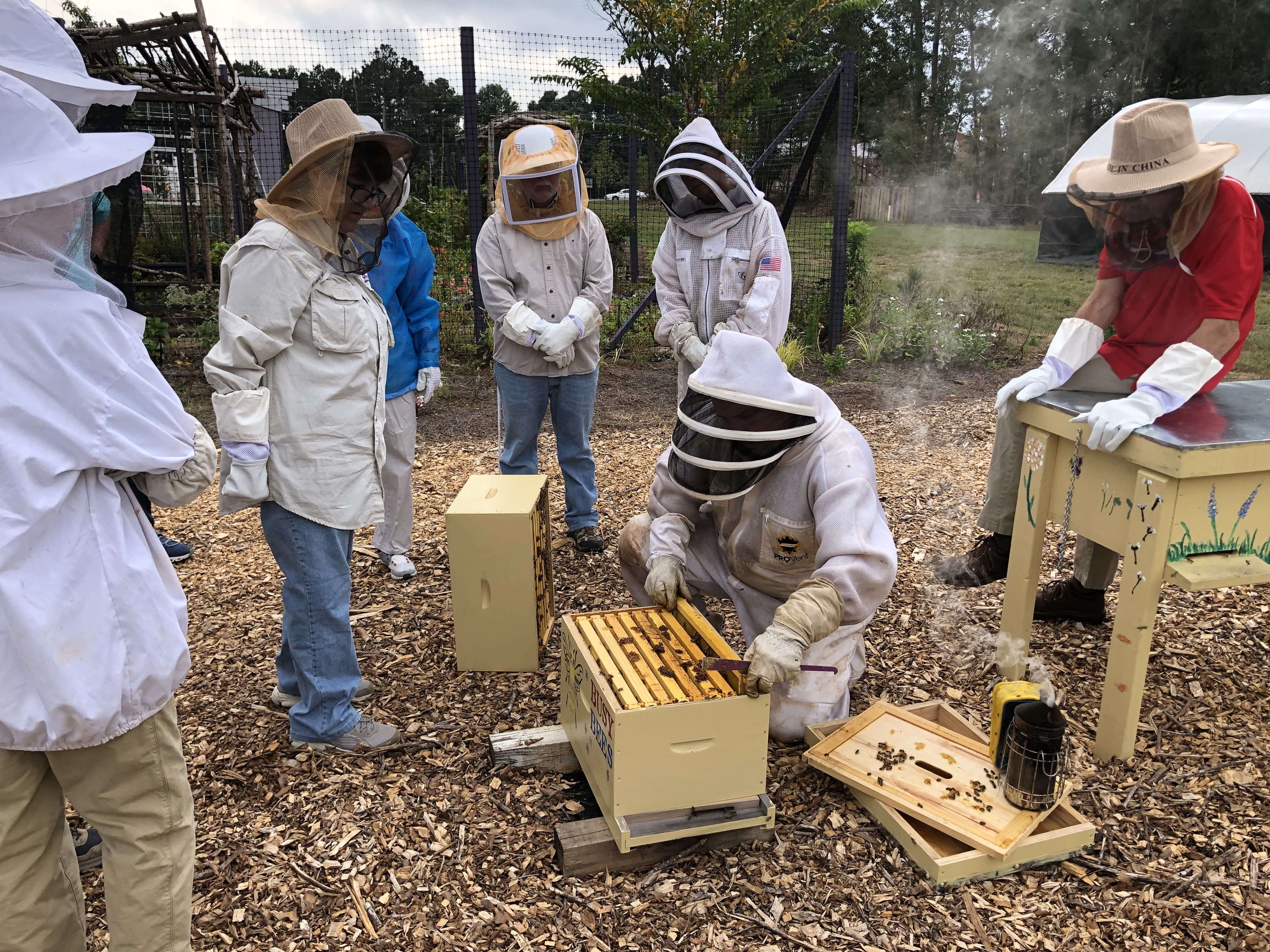 A group of budding bee keepers gather for an instructional hive check around bee boxes at the Clarke County Extension office. Smoke is visible in the background.