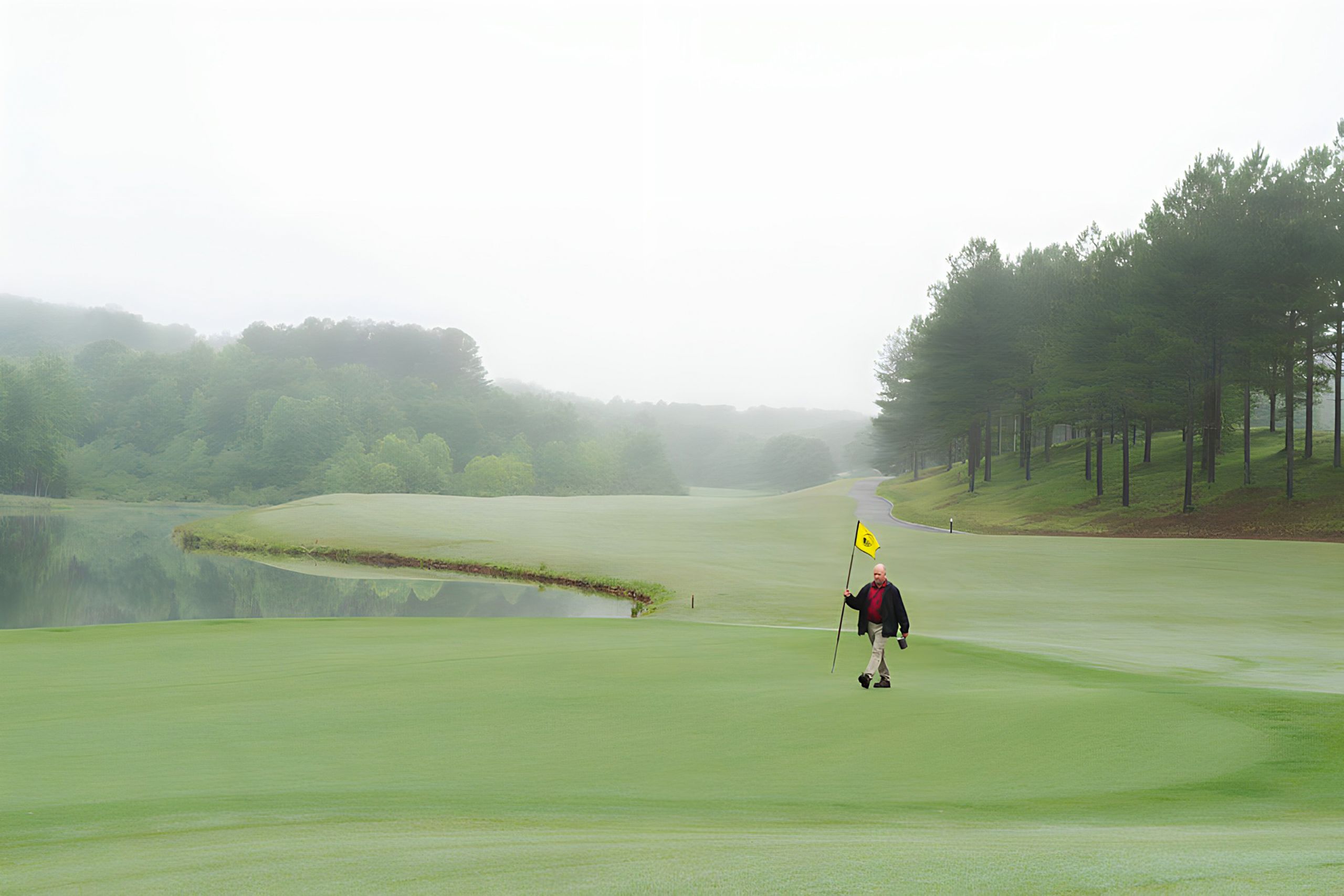 Jim Evans walks across a misty golf course, holding a yellow flag, surrounded by green fairways, a calm water, and tall trees.