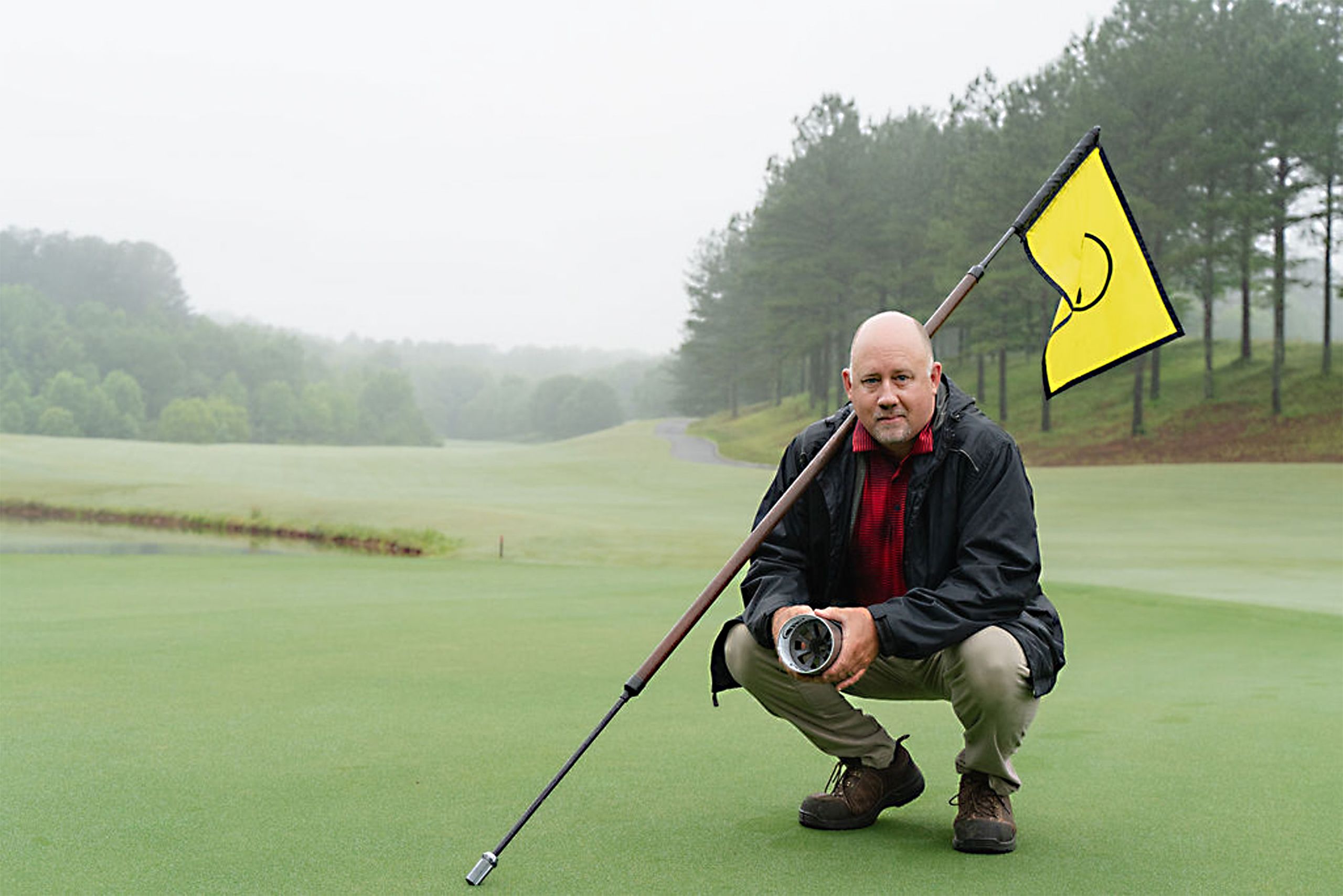 Jim Evans kneels on a golf course, holding an object, with a yellow flag in the background, set against a misty, serene landscape.