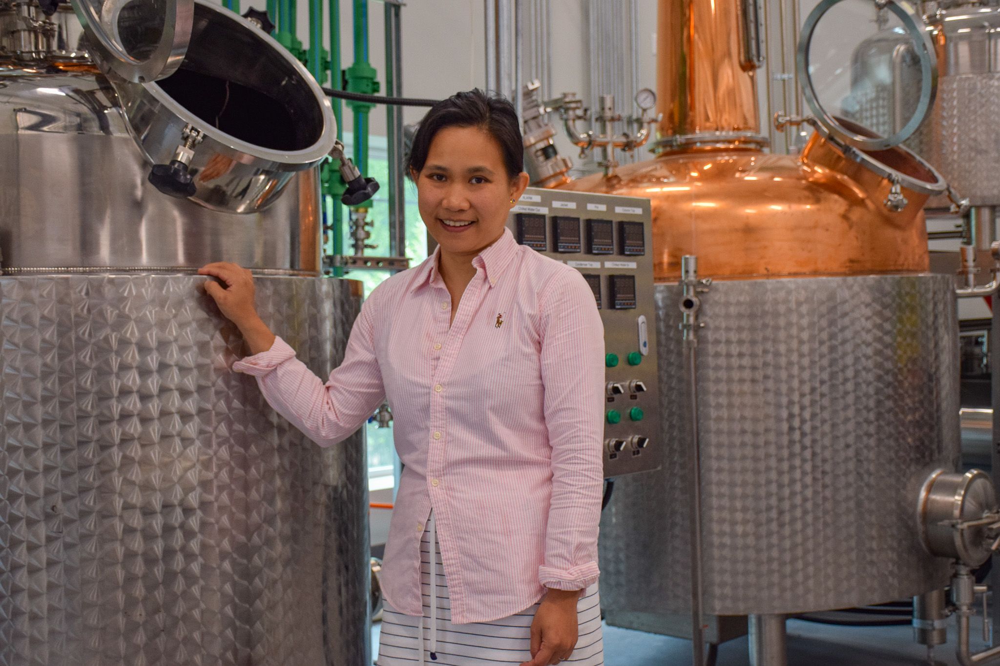 Supakana Nagachinta, owner and co-founder of OAK House Distillery stands next to metal distillery equipment. She wears a pale pink button up and a white striped skirt. 