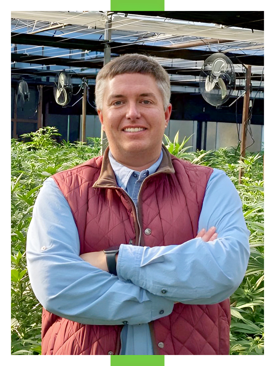 A male wearing a blue shirt and red vest stands in a greenhouse with his arms crossed.