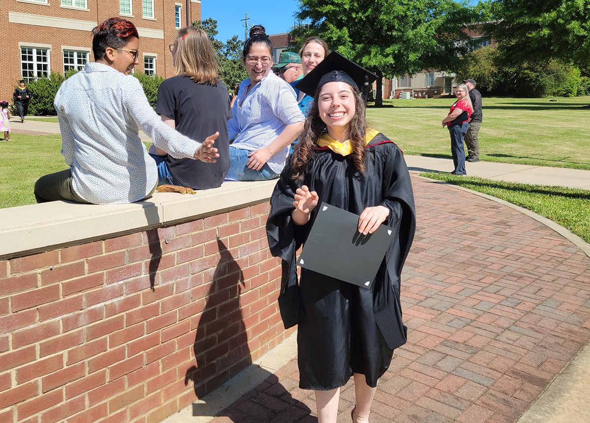 Raad poses in her cap and gown while friends congratulate her from the background