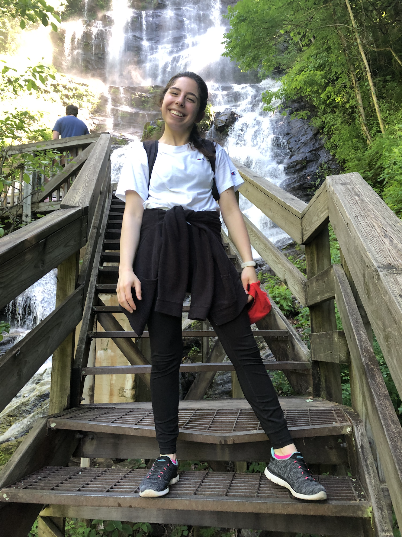 Raad smiles while on a set of stairs during a waterfall hike. A dramatic waterfall and another hiker are visible behind her