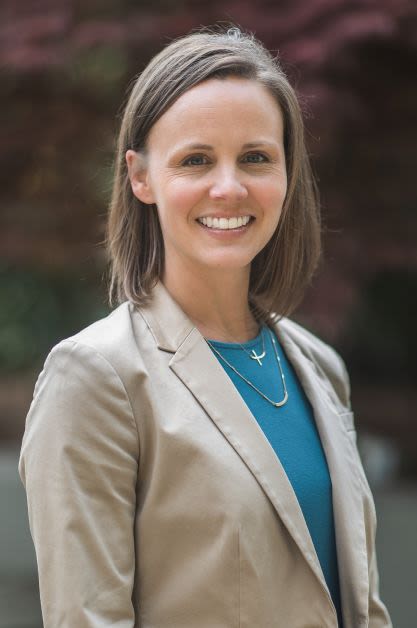 A smiling woman wearing a blue top and tan blazer faces the camera in an outdoor setting.