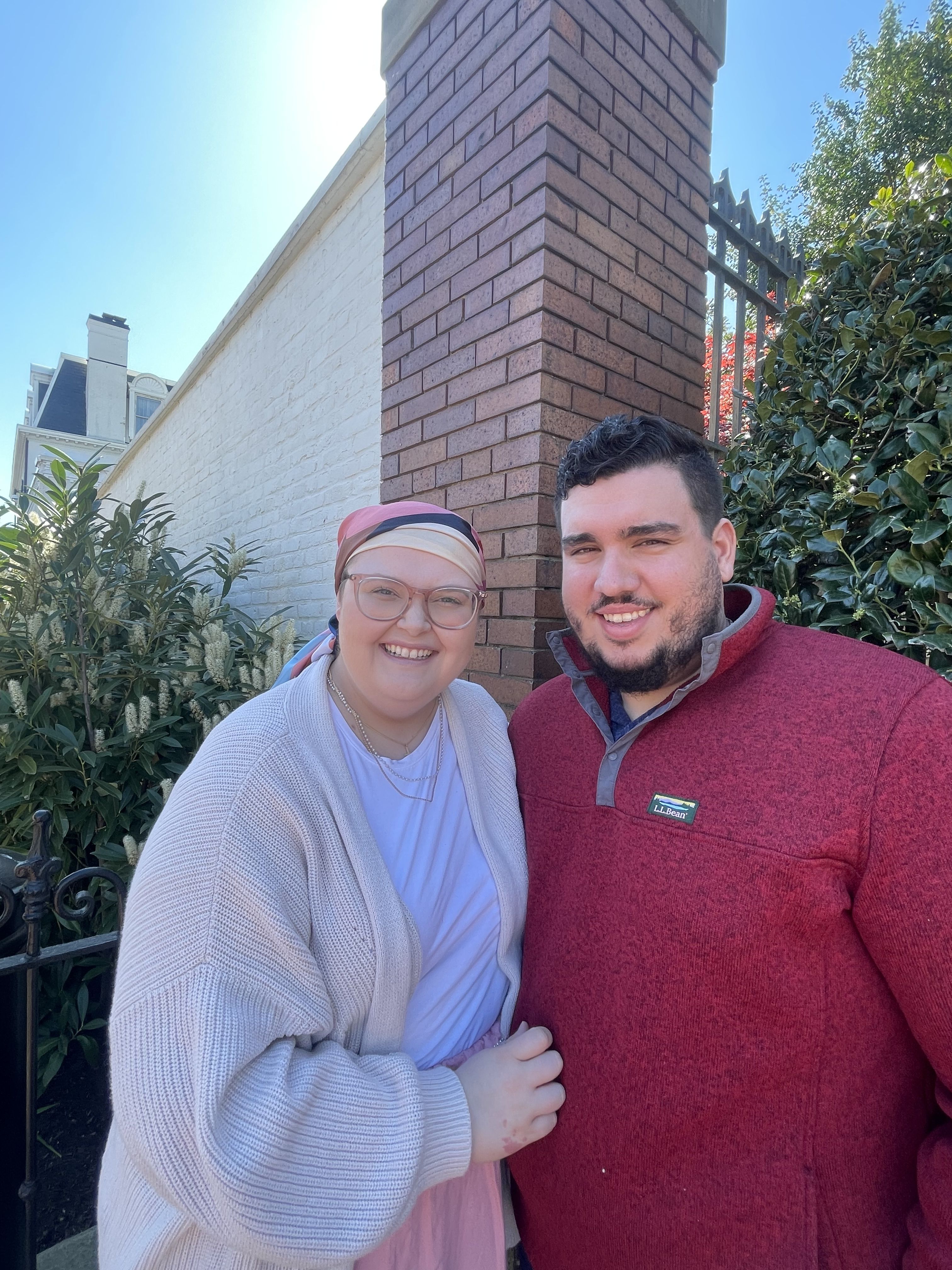 A smiling man in a red jacket and a smiling woman in a light sweater and headscarf stand together outside.