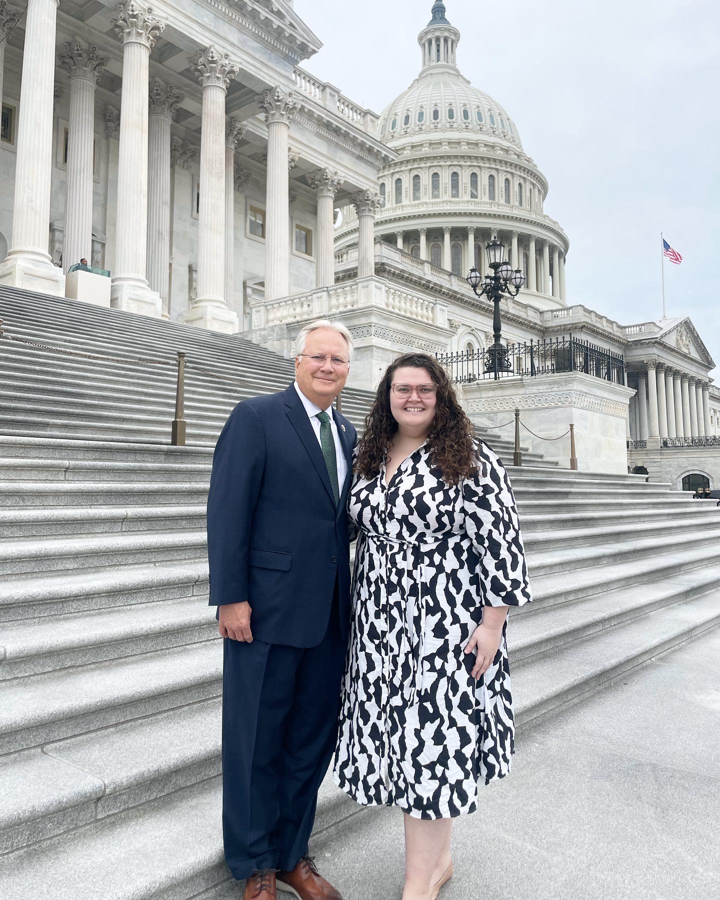A man in a suit and a woman in a black-and-white dress stand on the steps of a government building, with the U.S. Capitol in the background.