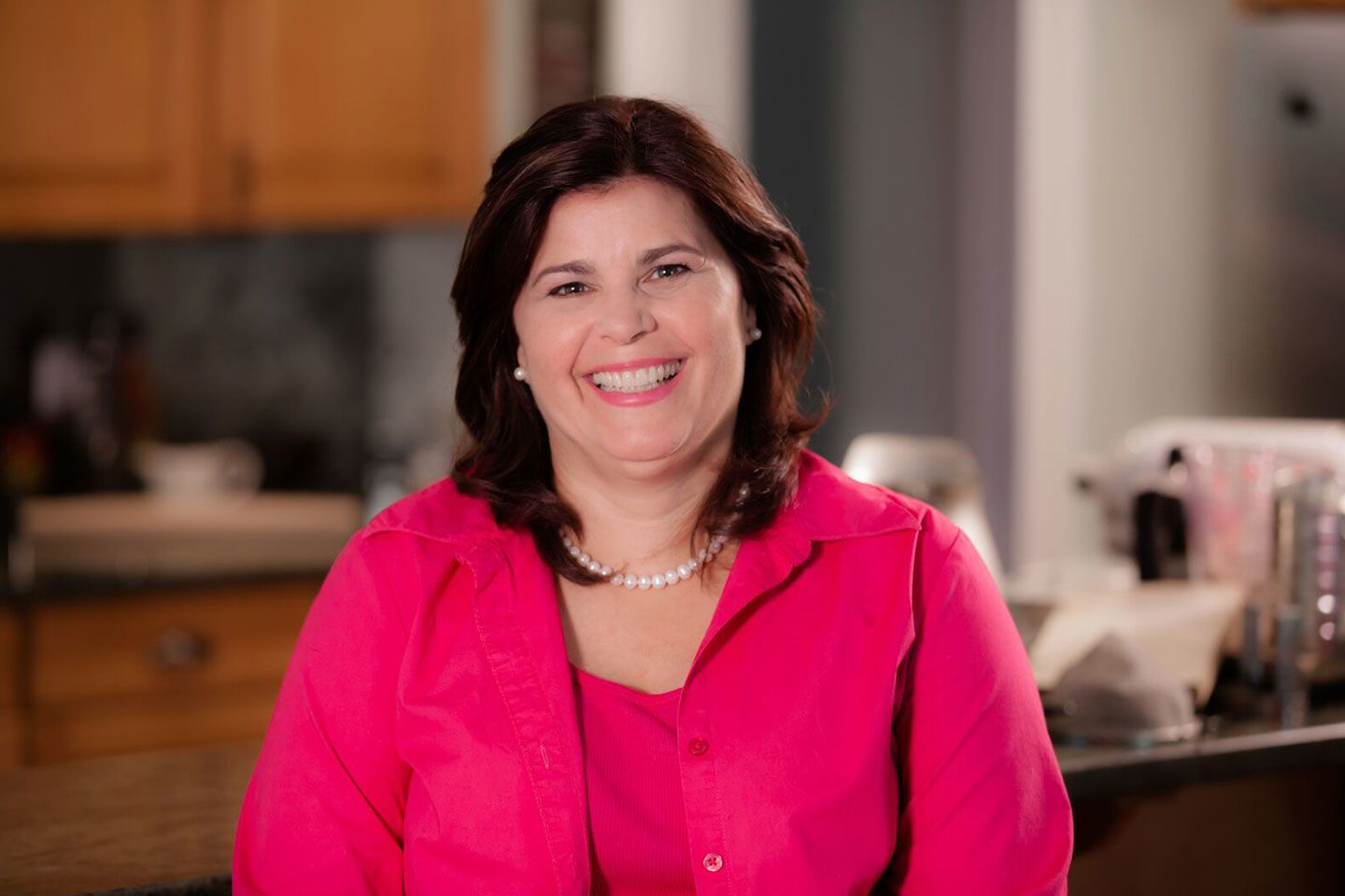 Shona Jonson sits in her kitchen wearing a bright pink blouse and matching jacket
