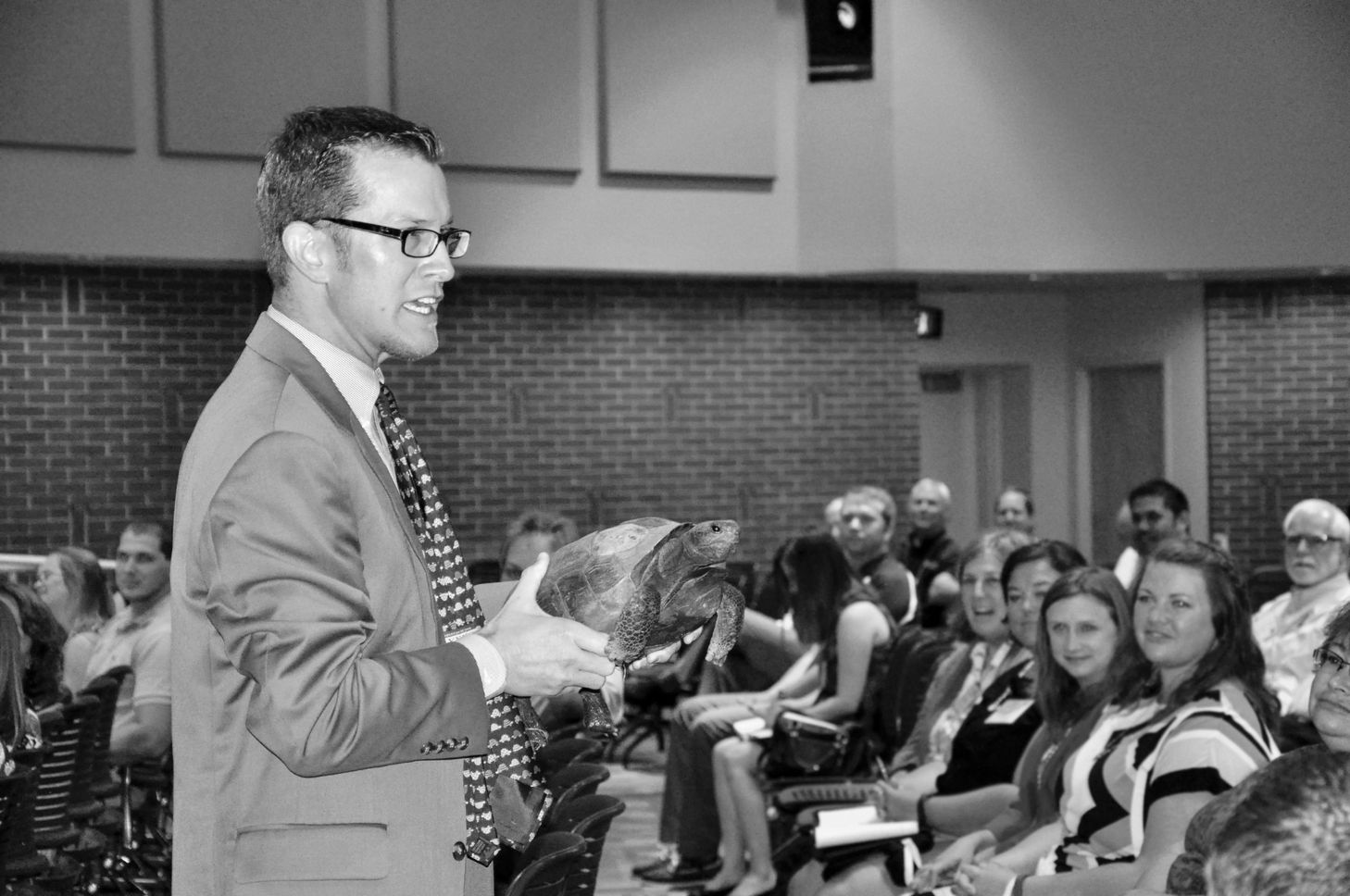 In black and white, Fuhrman, wearing a tie covered in turtles, holds up a turtle while giving a presentation to adults in an auditorium