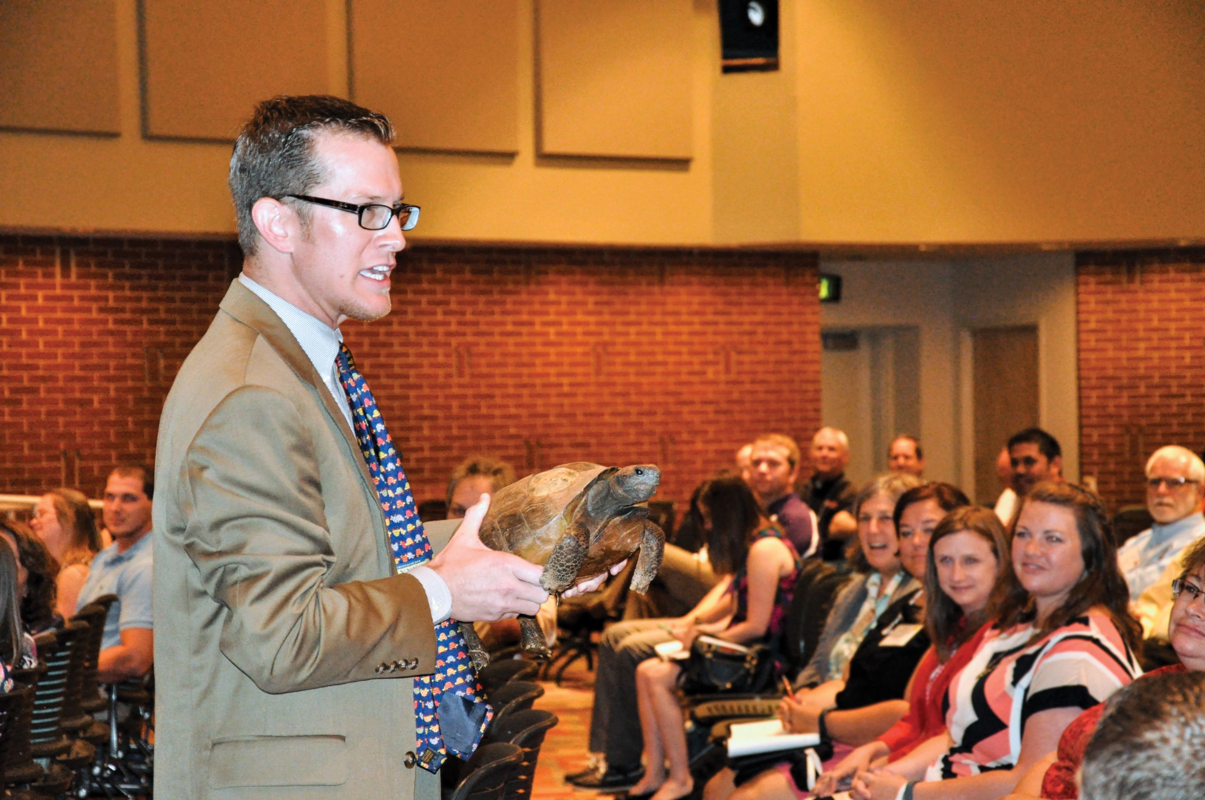 In full color, Fuhrman, wearing a tie covered in turtles, holds up a turtle while giving a presentation to adults in an auditorium