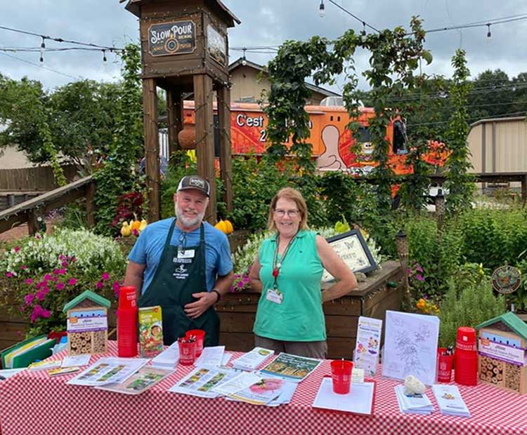 A man and a woman stand behind a table with a red and white checkered tablecloth. The table is covered with various papers, brochures and giveaway items. The two people are standing in front of a garden with a wooden tower and an orange food truck in the background. 