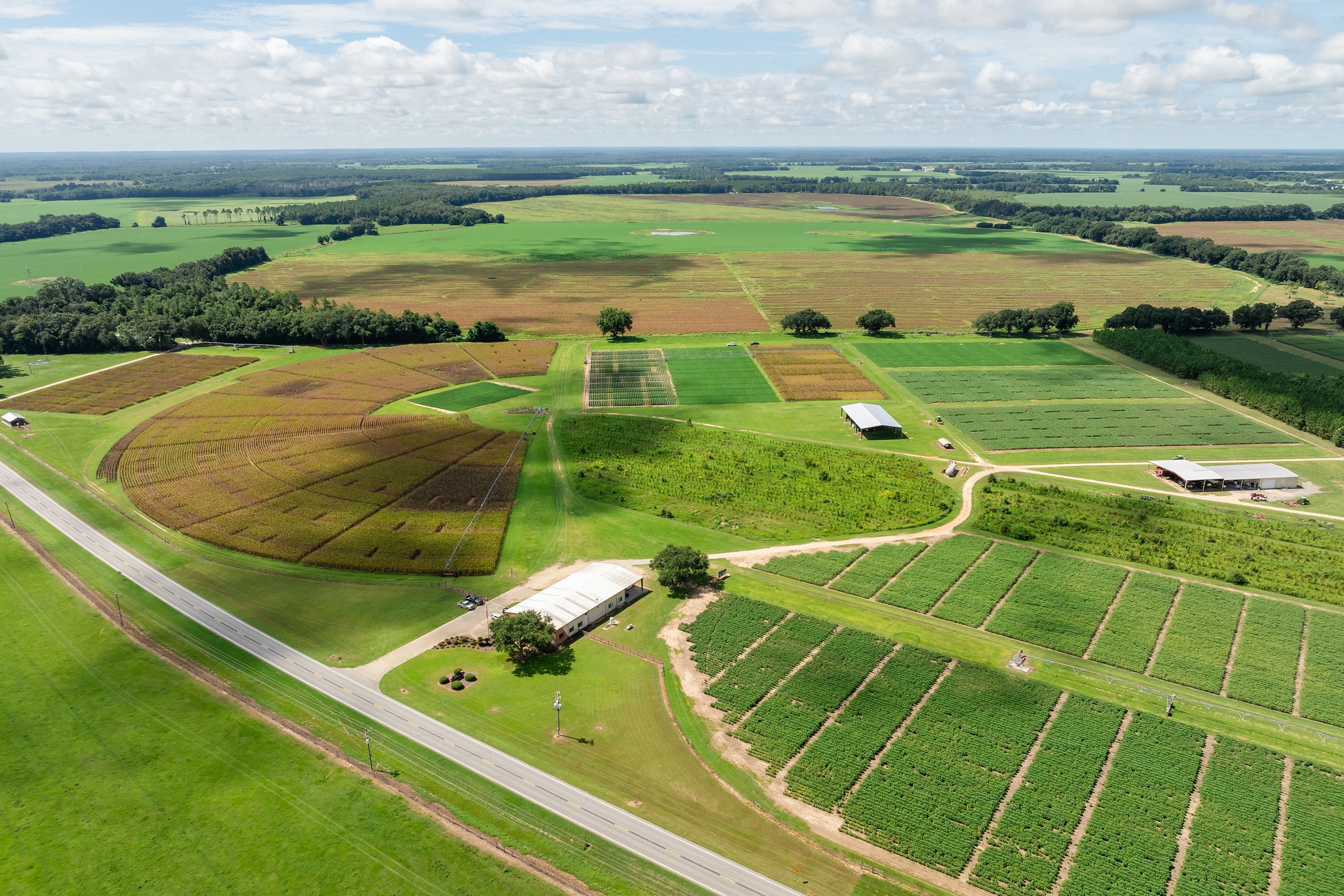 An aerial view of a sprawling agricultural landscape featuring neatly organized fields, patches of greenery, and farm buildings, all under a partly cloudy sky.