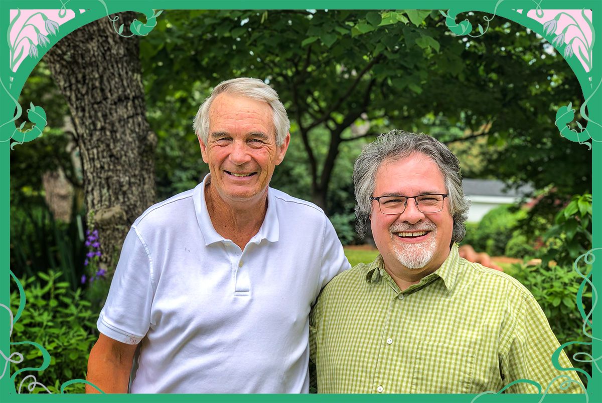 Two smiling men stand outdoors in front of leafy green trees, one wearing a white polo shirt and the other wearing glasses and a green checkered shirt.