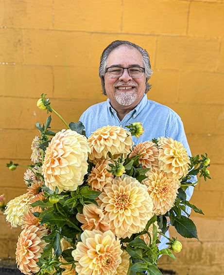 A man smiling while holding a bouquet of yellow dahlias against a bright yellow wall.