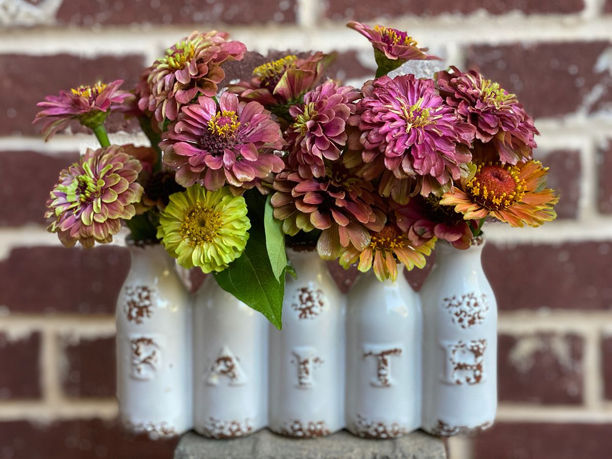 Bouquets of pink, purple, and yellow zinnias arranged in a white ceramic vase with the word "EARTH" embossed on its bottles, set against a red brick wall.