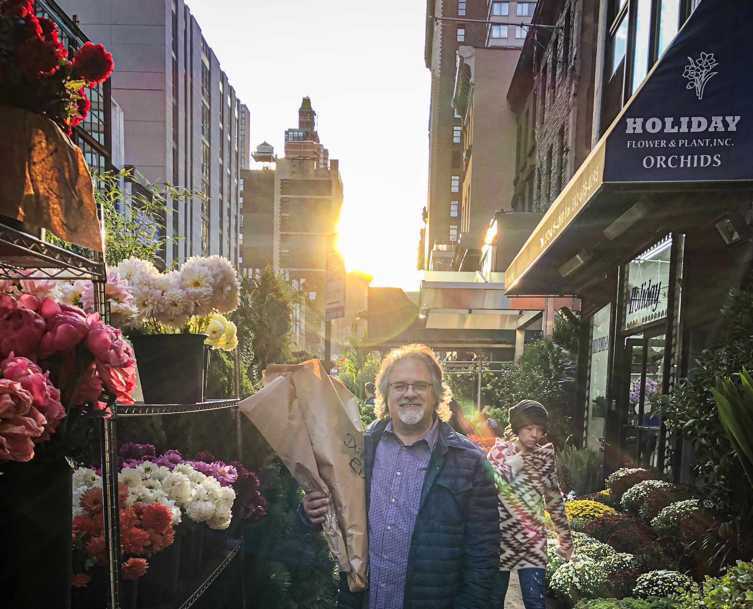 Man smiling with flowers at a market, holding a bag, sunlight glowing behind him.