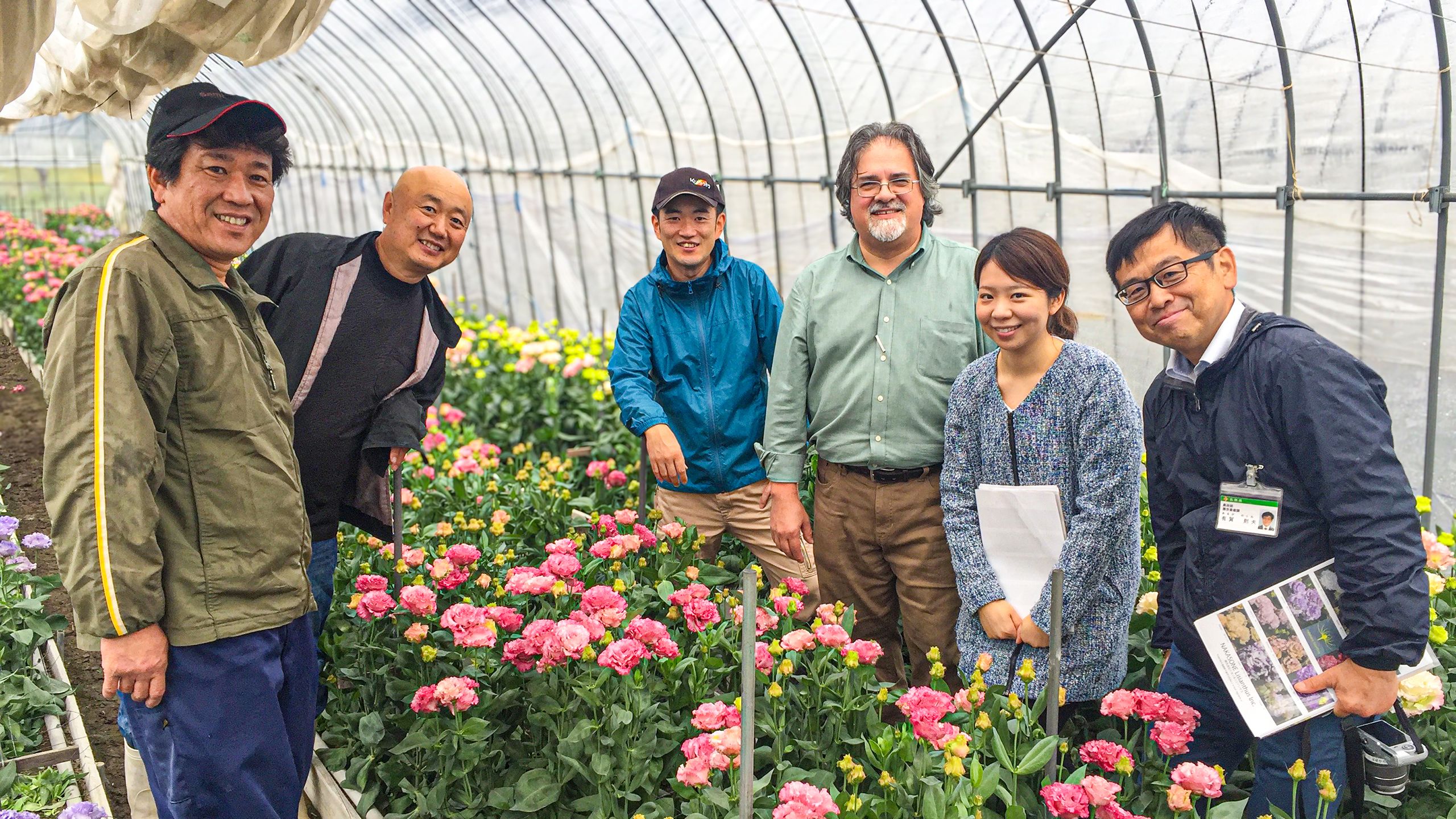 Six people stand and smile among rows of blooming pink and yellow flowers inside a greenhouse. Some hold papers, and all appear to be posing for the photo.